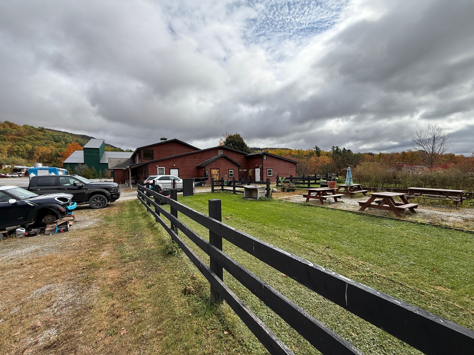 Rustic lodge with picnic tables, parked cars, and a black fence under cloudy autumn skies
