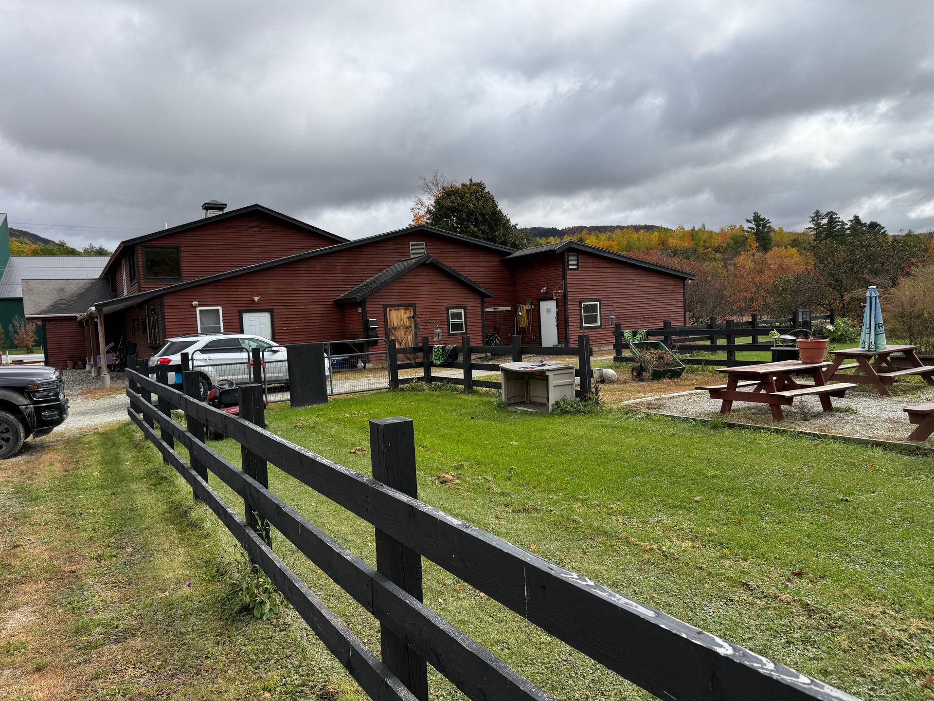 Rustic red lodge with picnic tables, a black fence, parked vehicles, and cloudy sky over a grassy yard