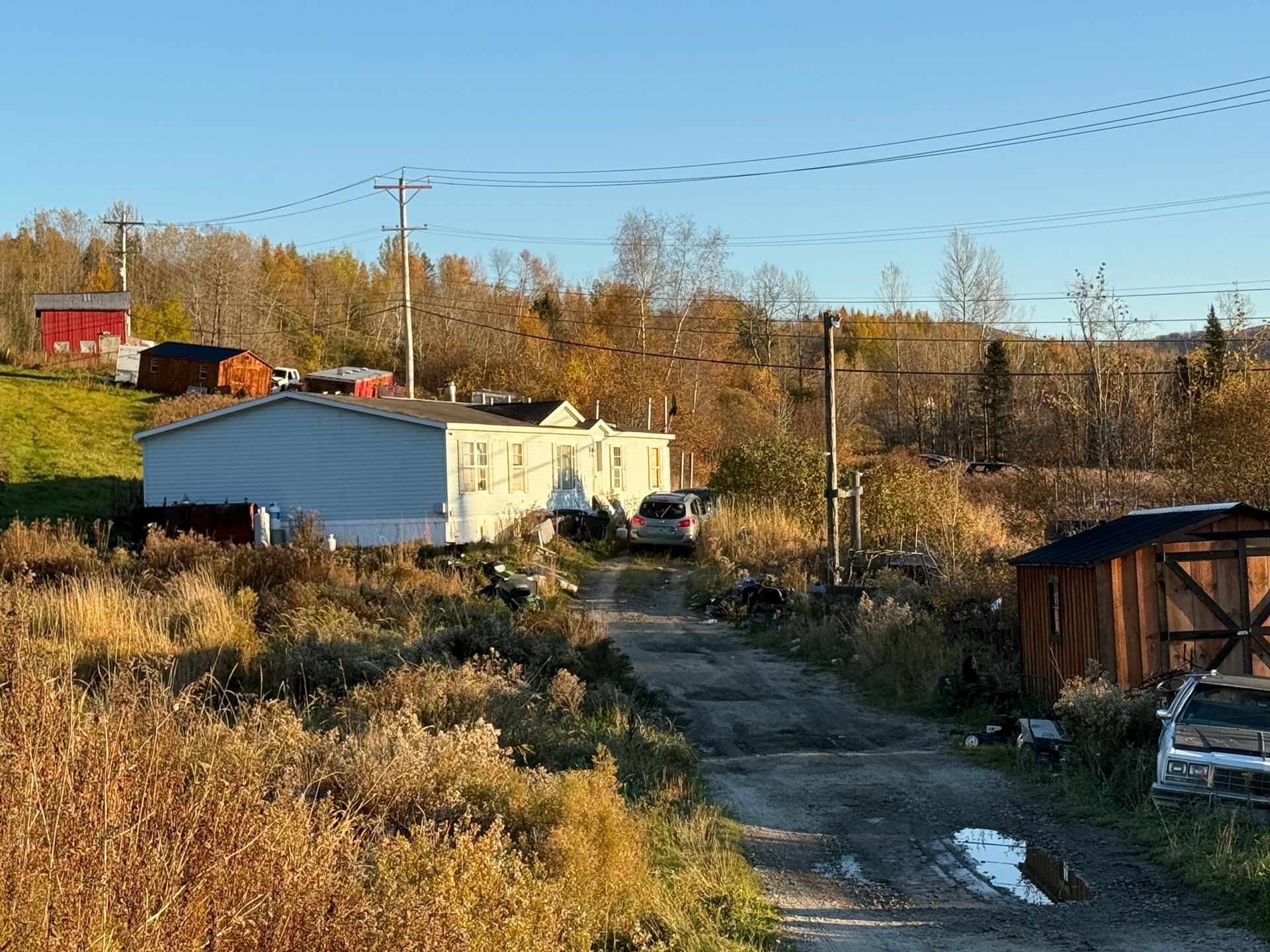 Rural dirt road past a white trailer and sheds in autumn fields under a clear blue sky