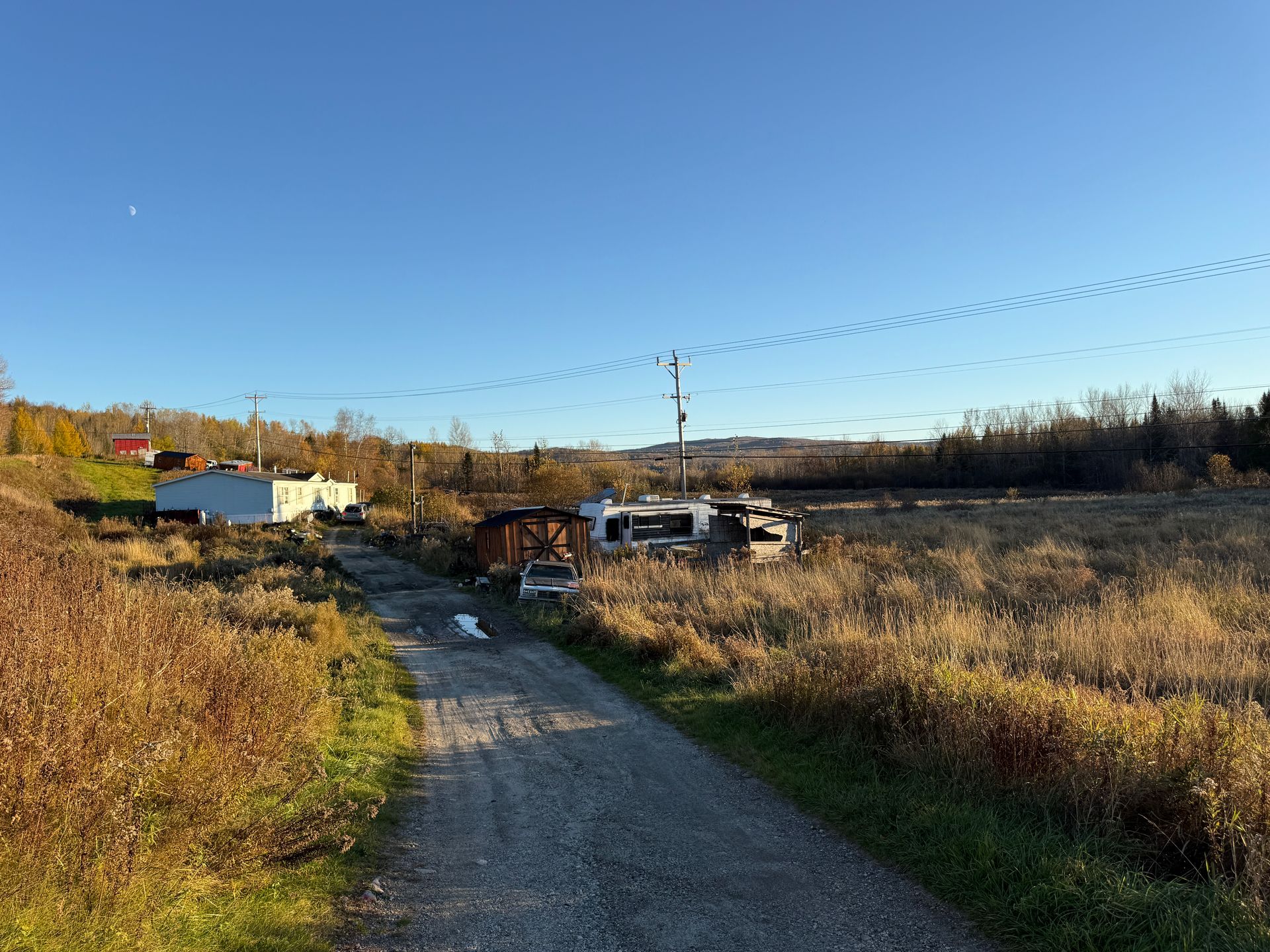 Rural gravel road beside fields and houses under a clear blue sky at sunset