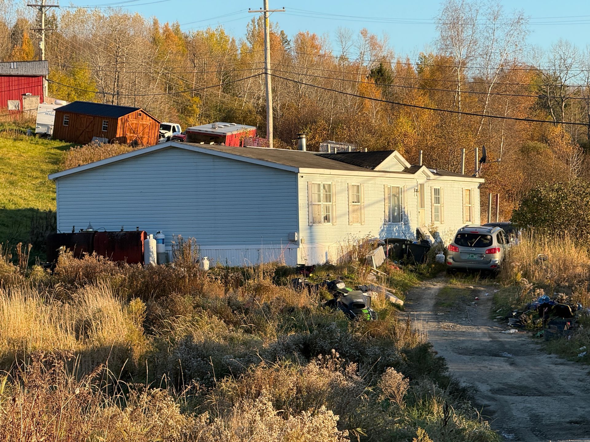 Mobile homes along a dirt driveway in a rural autumn setting, with parked cars and trees in the background