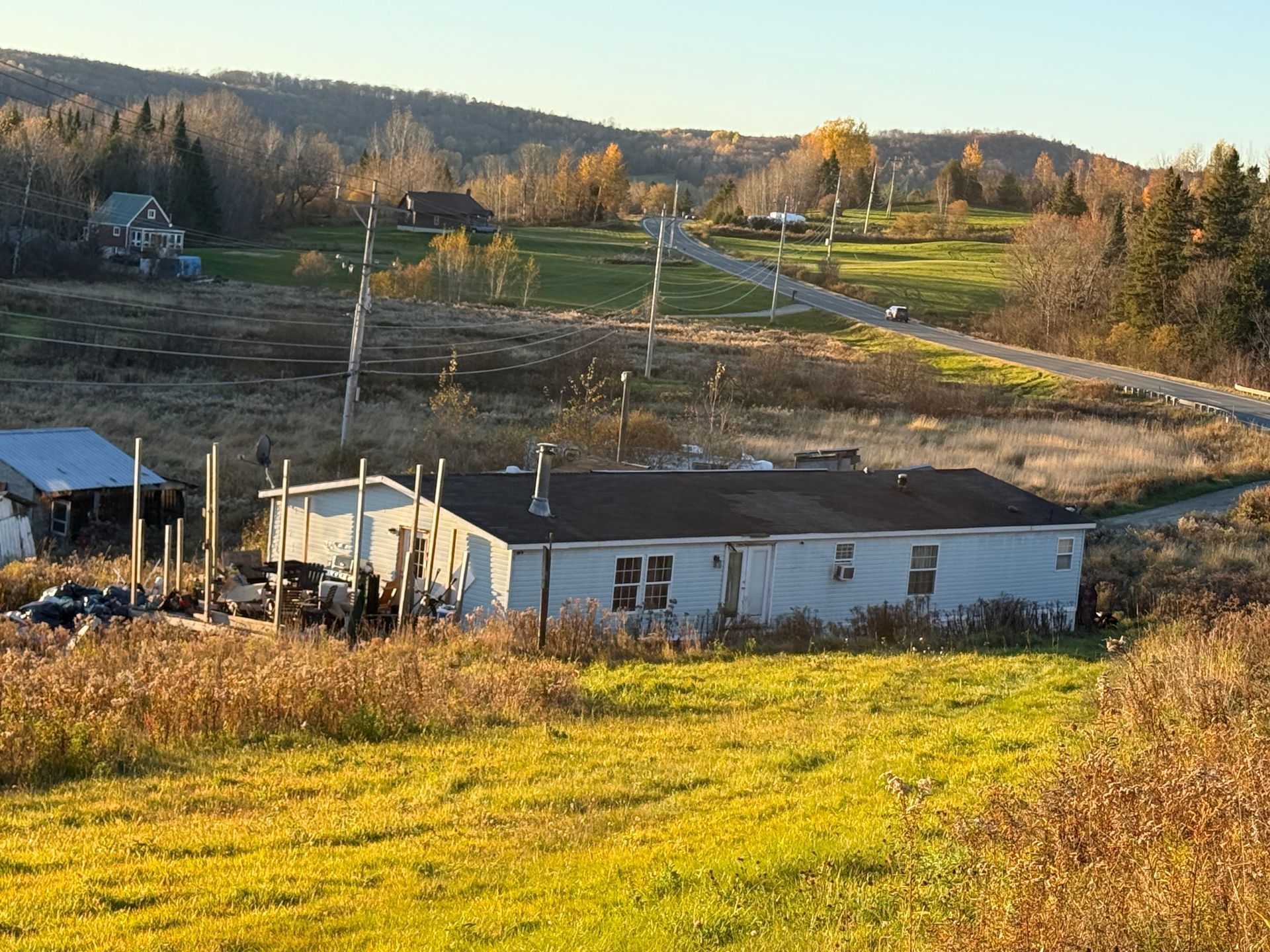 Rural hillside with a small white house, dirt roads, power lines, and sunlit fields at sunset