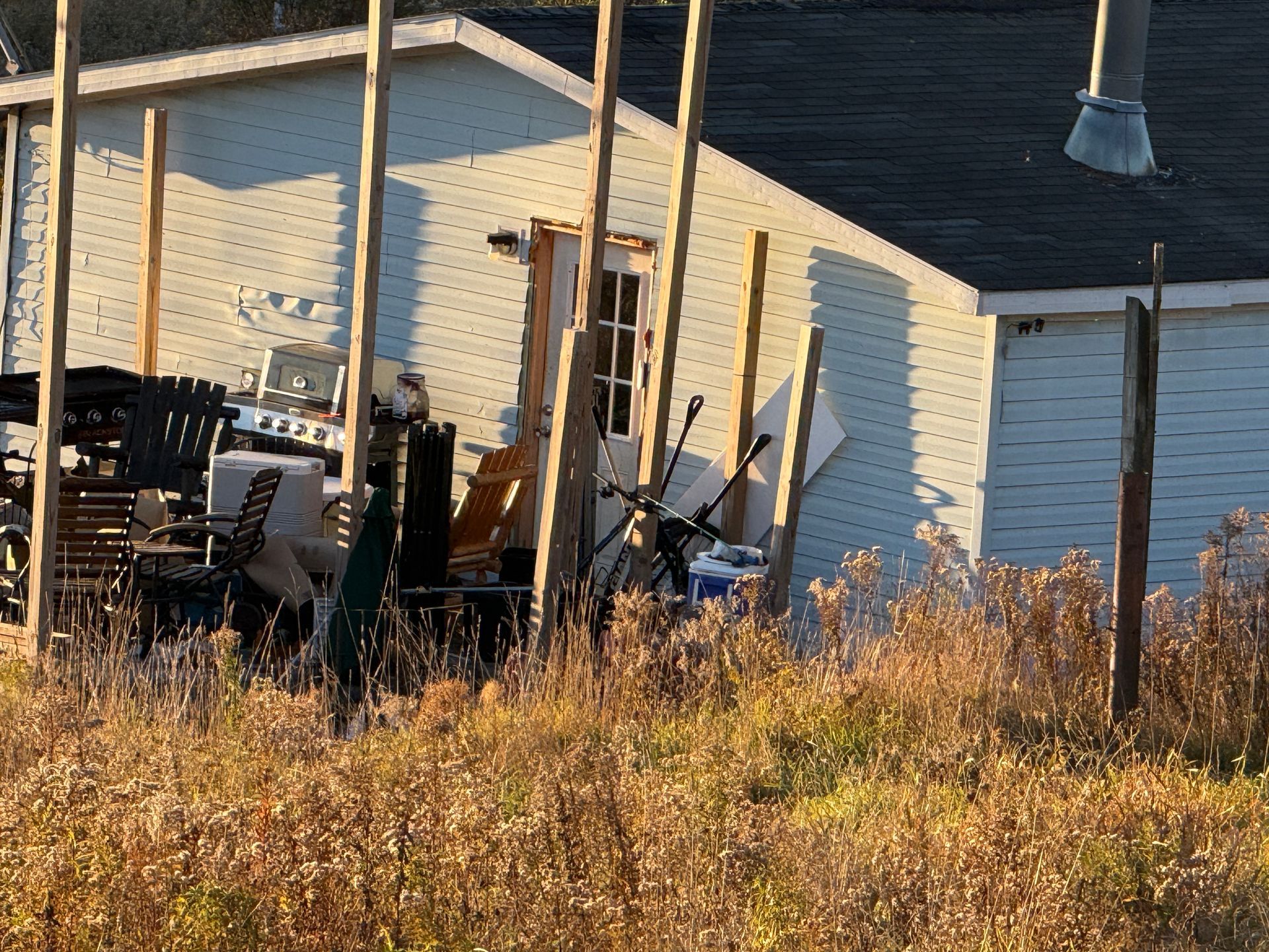 Backyard with a white shed, stacked chairs, and tools beside tall dry grass