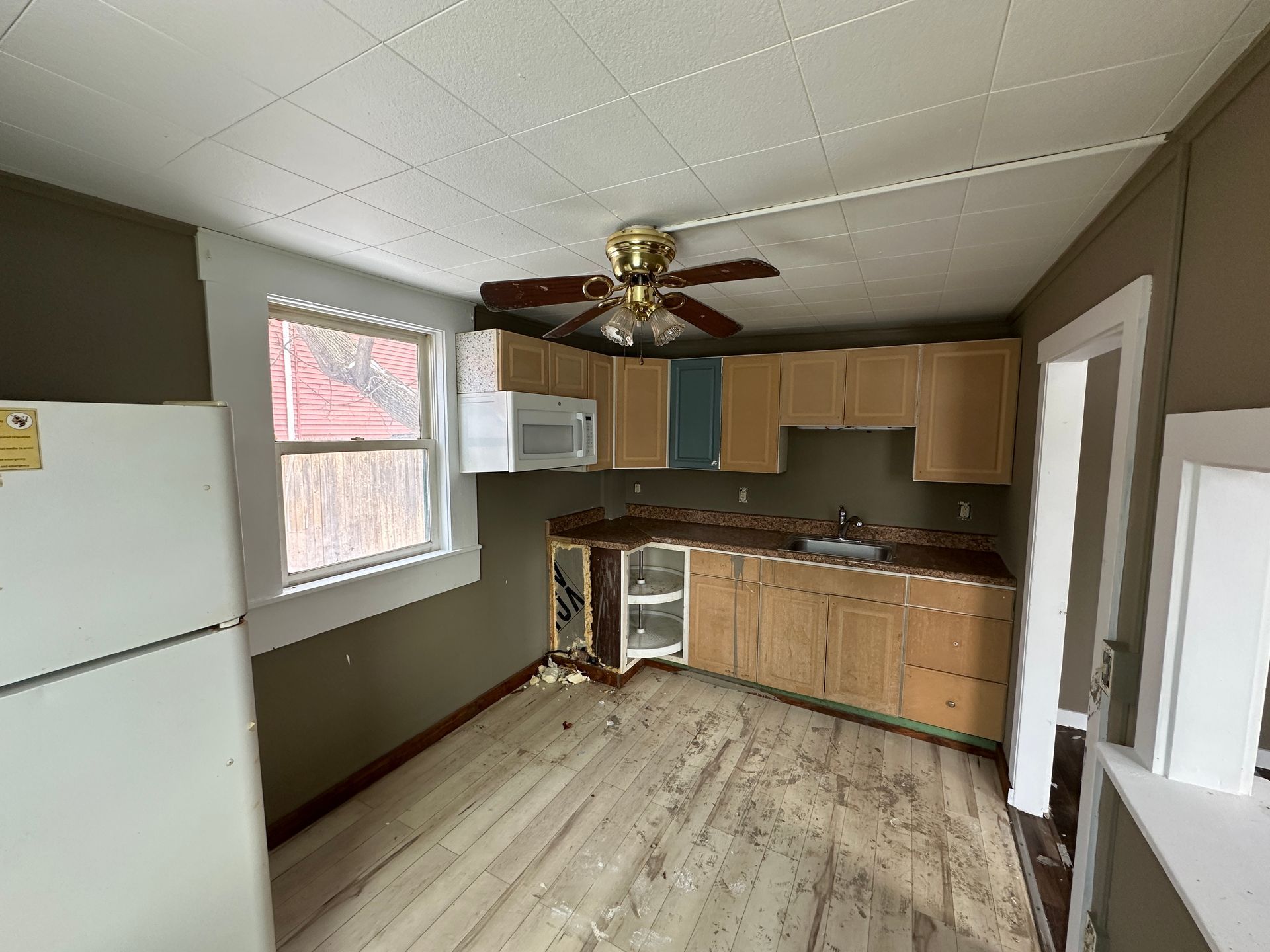 Kitchen with light wood cabinets, floor, and window. Refrigerator on the left, sink and microwave.