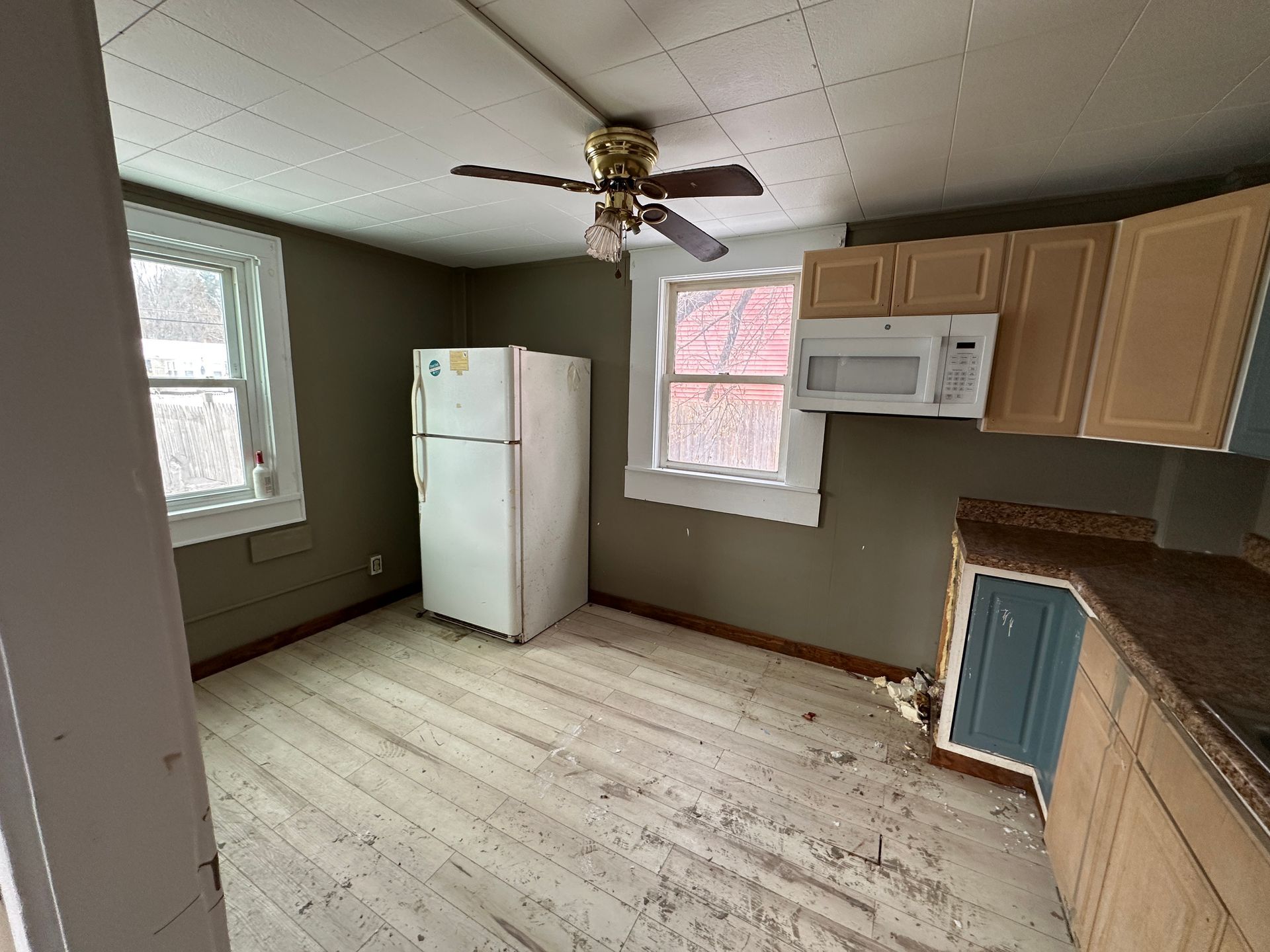 Kitchen interior with fridge, cabinets, microwave, windows, ceiling fan, and flooring.