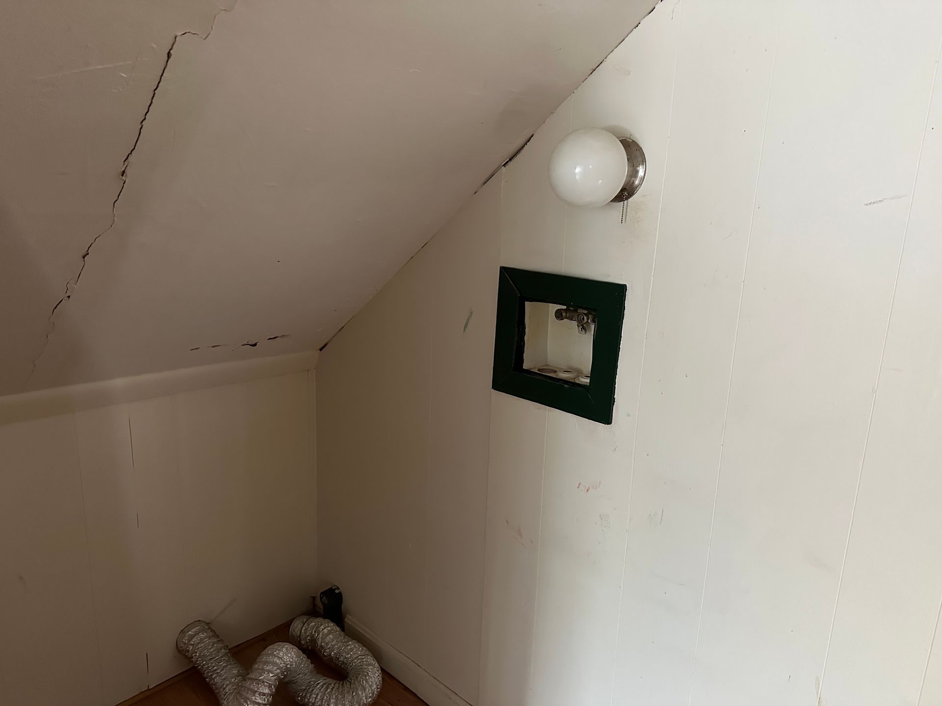 Small, white-painted room with sloped ceiling, light fixture, vent, and exposed dryer vent hose.