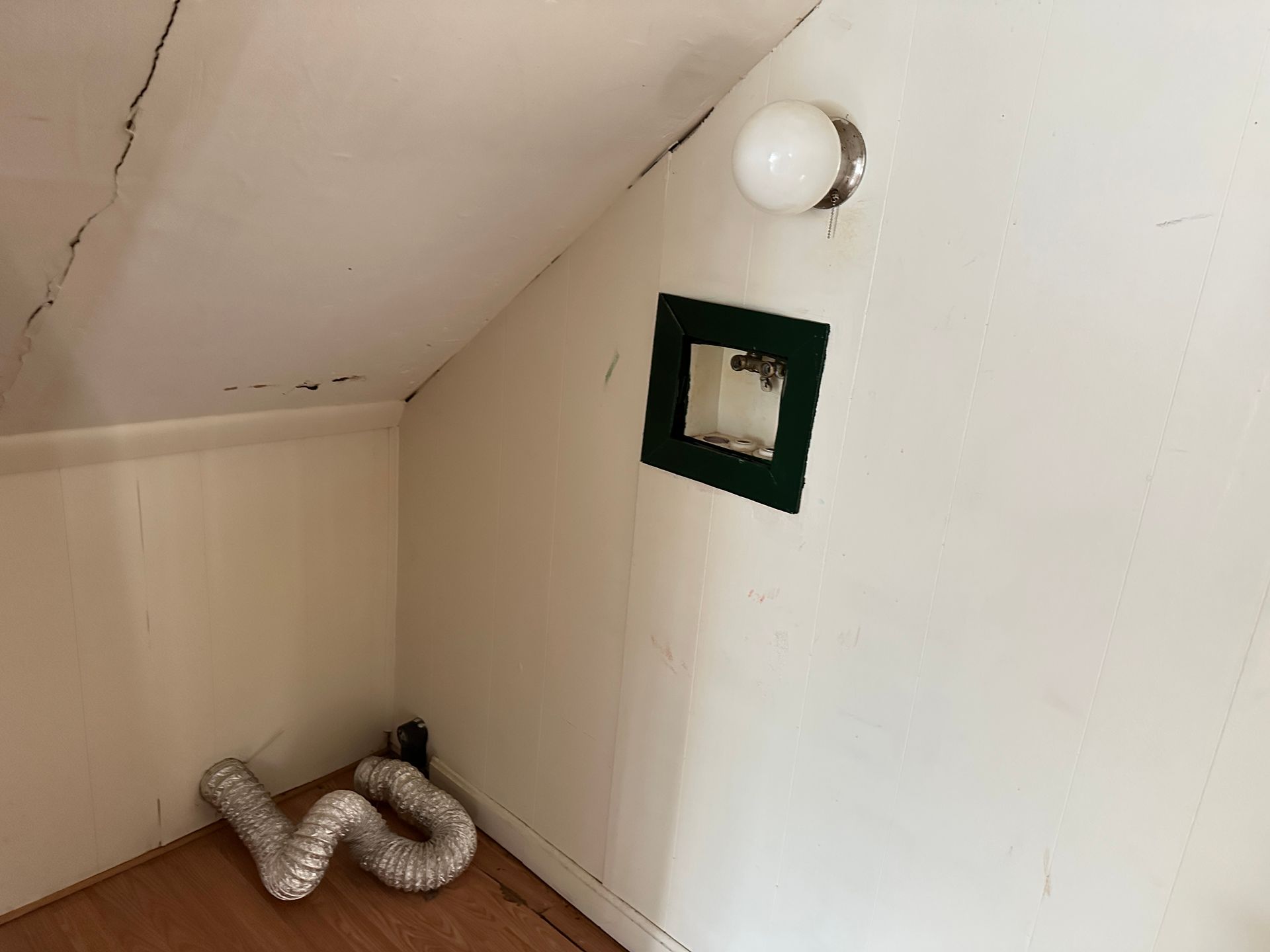 Sloping attic room with white walls, vent pipe, small window, and light fixture.
