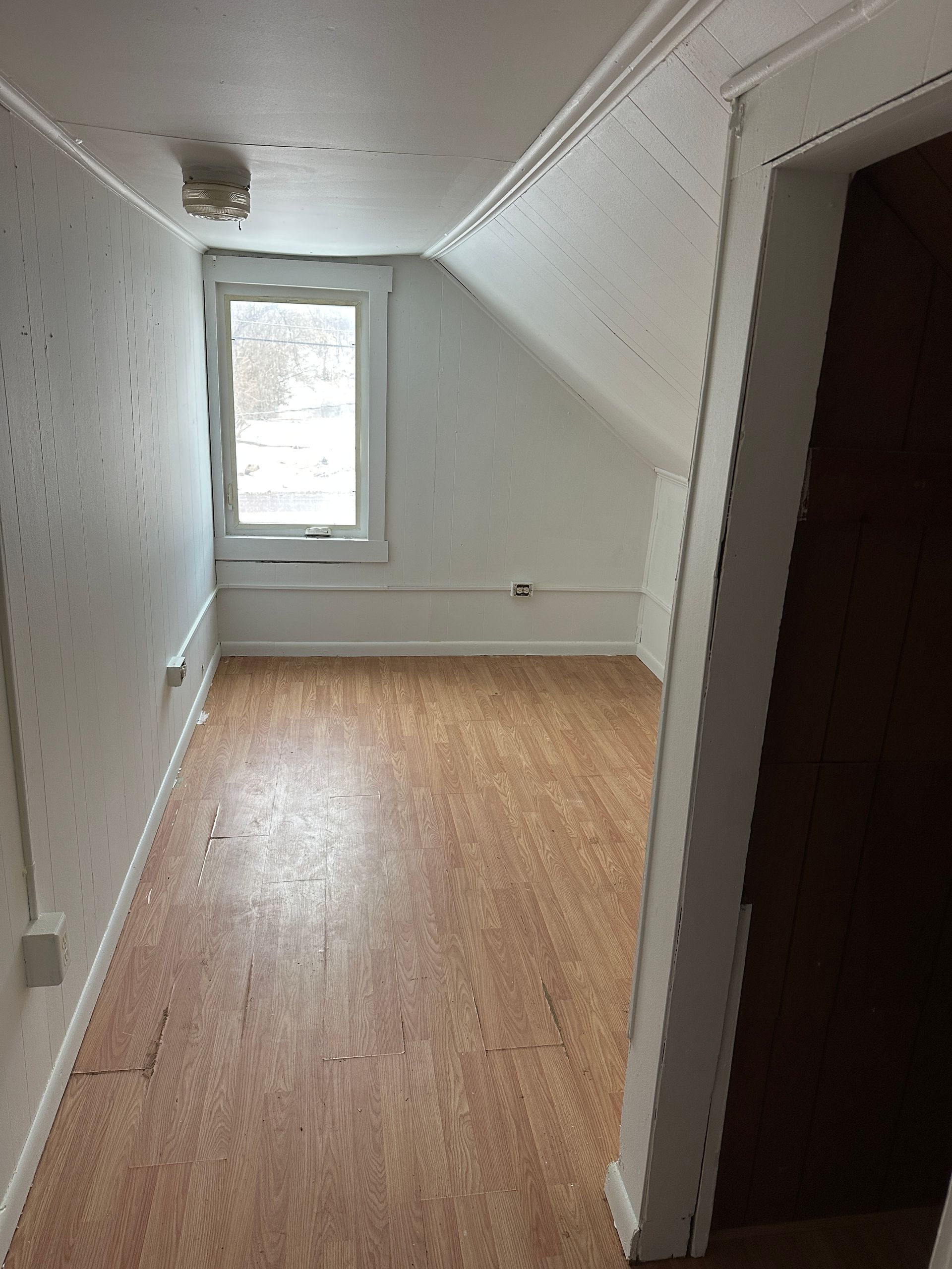 Attic room with window, white walls, angled ceiling, and wood-look flooring.