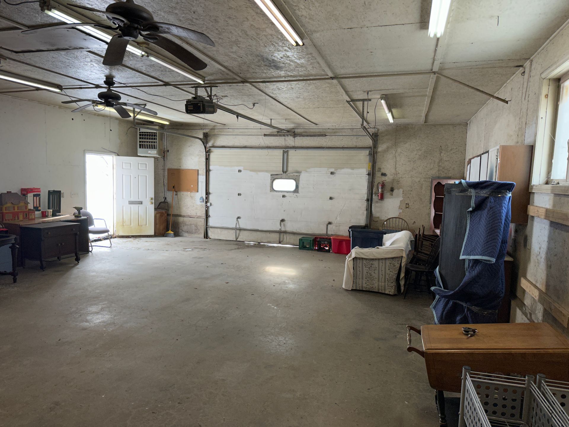 Empty garage interior with open door, cement floor, overhead lights, and furniture.
