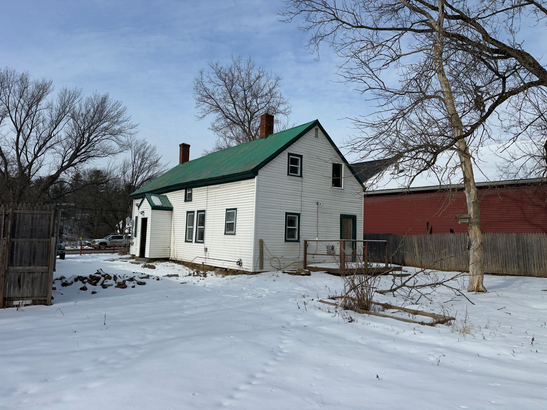 White two-story house with green roof, set in a snowy yard with a red barn visible in the background.