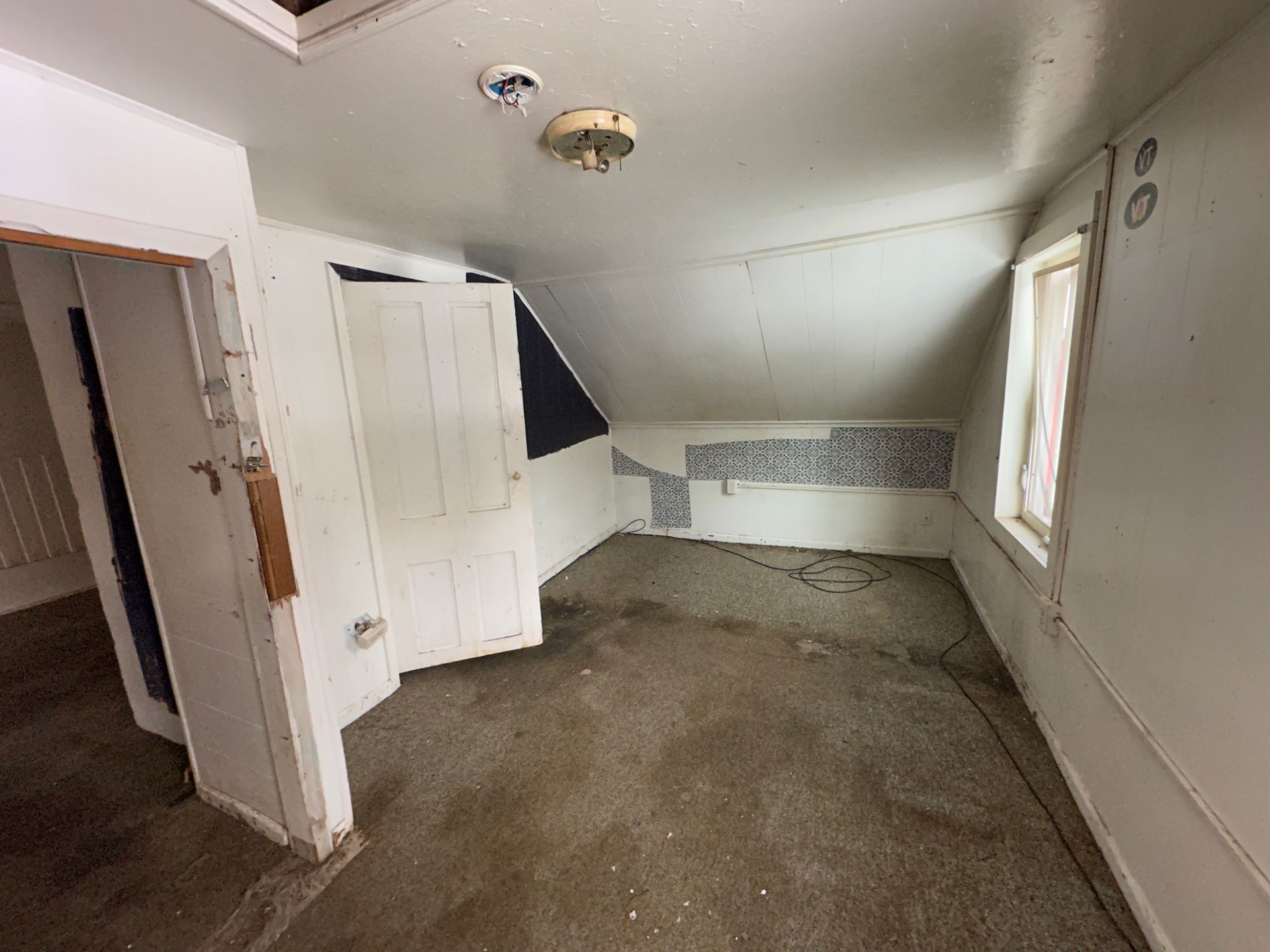 An attic room with a sloped ceiling, plain white walls, a single window, and a white wooden door leading to another area.