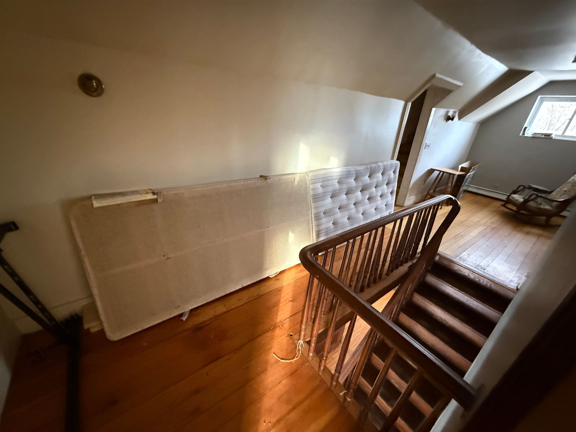 Bed frame and upholstered headboard against a slanted wall, top of staircase in the foreground, hardwood floors.