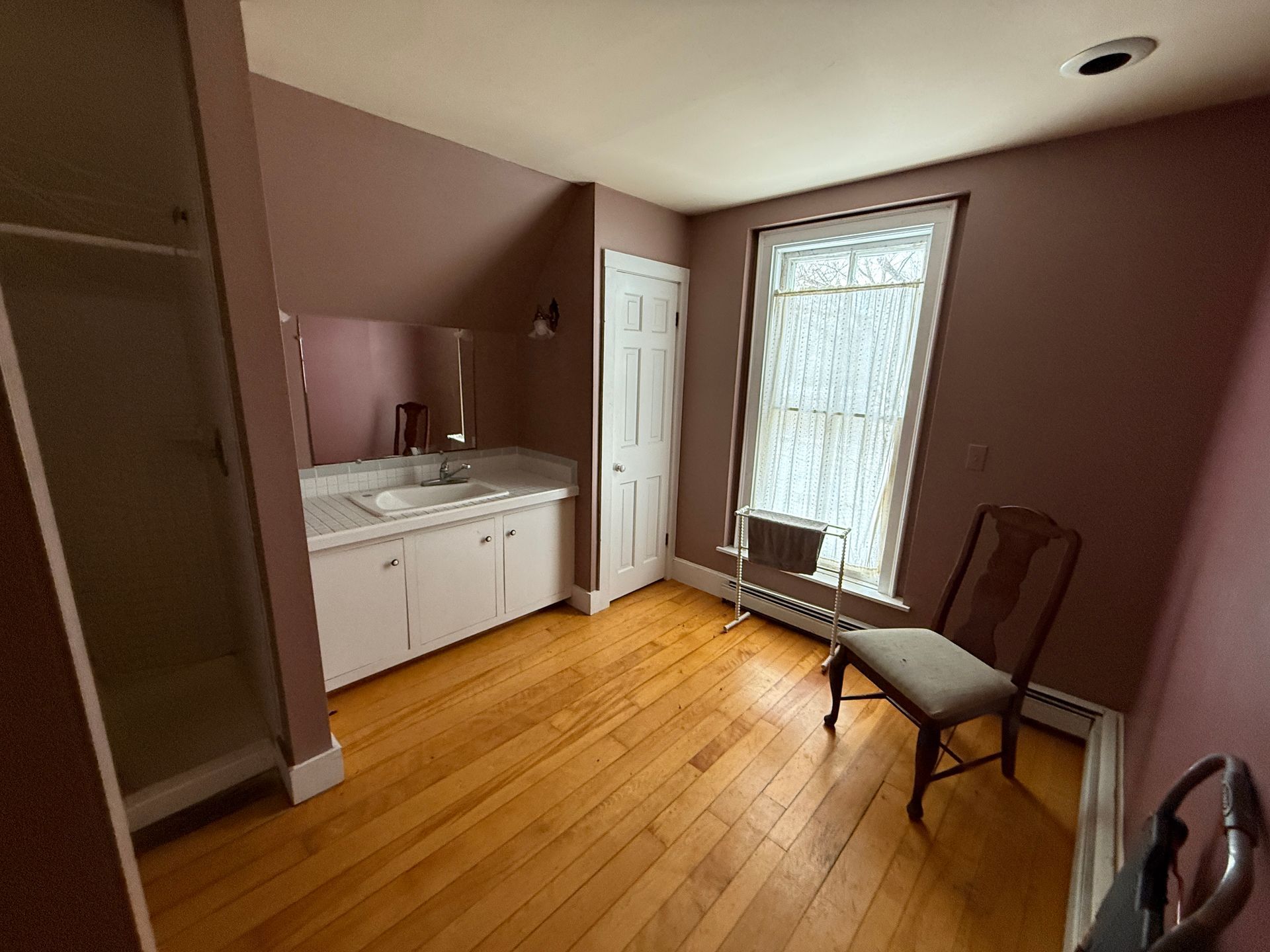 Bathroom with hardwood floor, vanity, white door, window, and a chair; mauve walls.