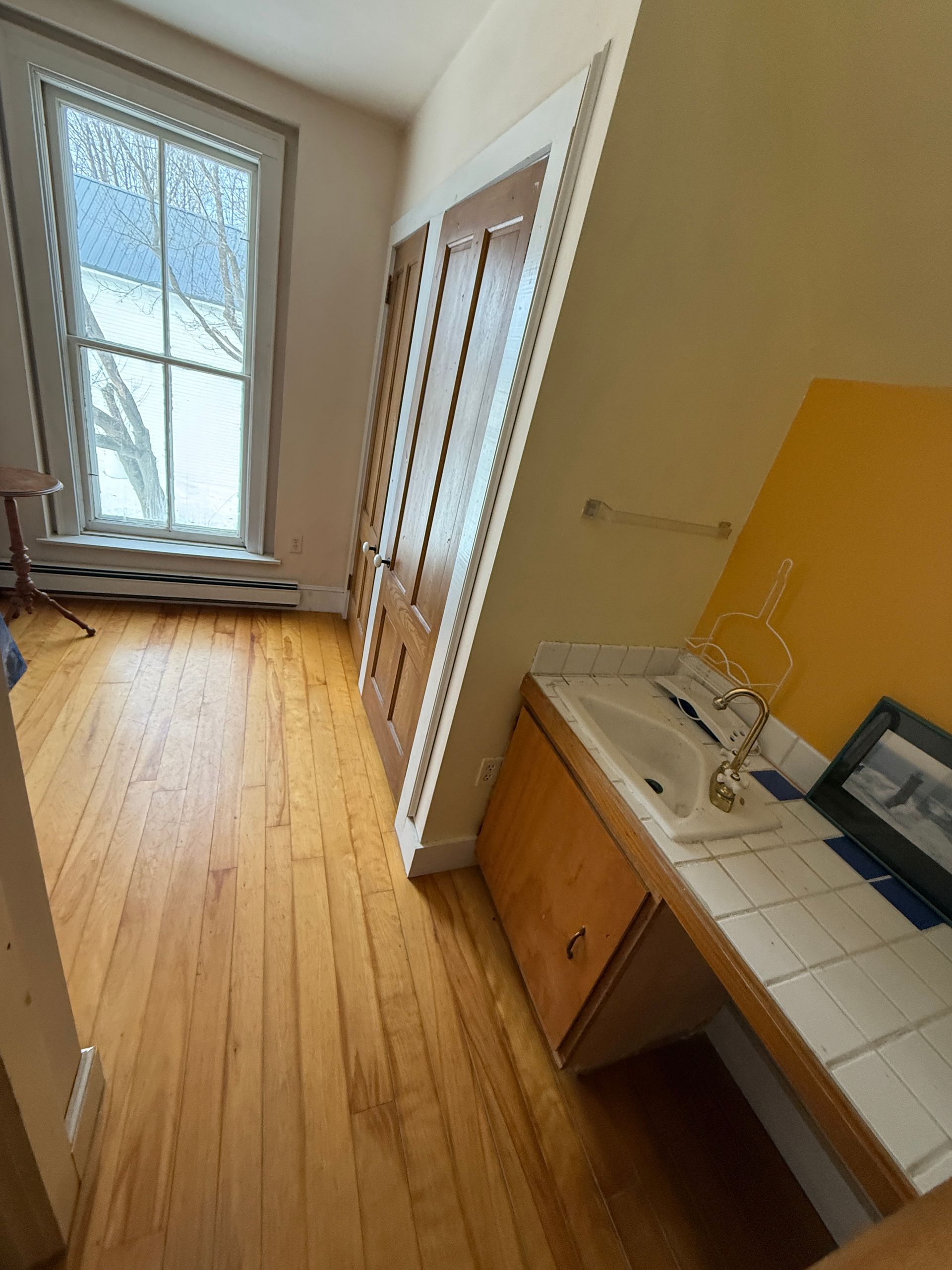 Wooden-floored room with a window, doorway, and sink. The walls are yellow, and there is a radiator.