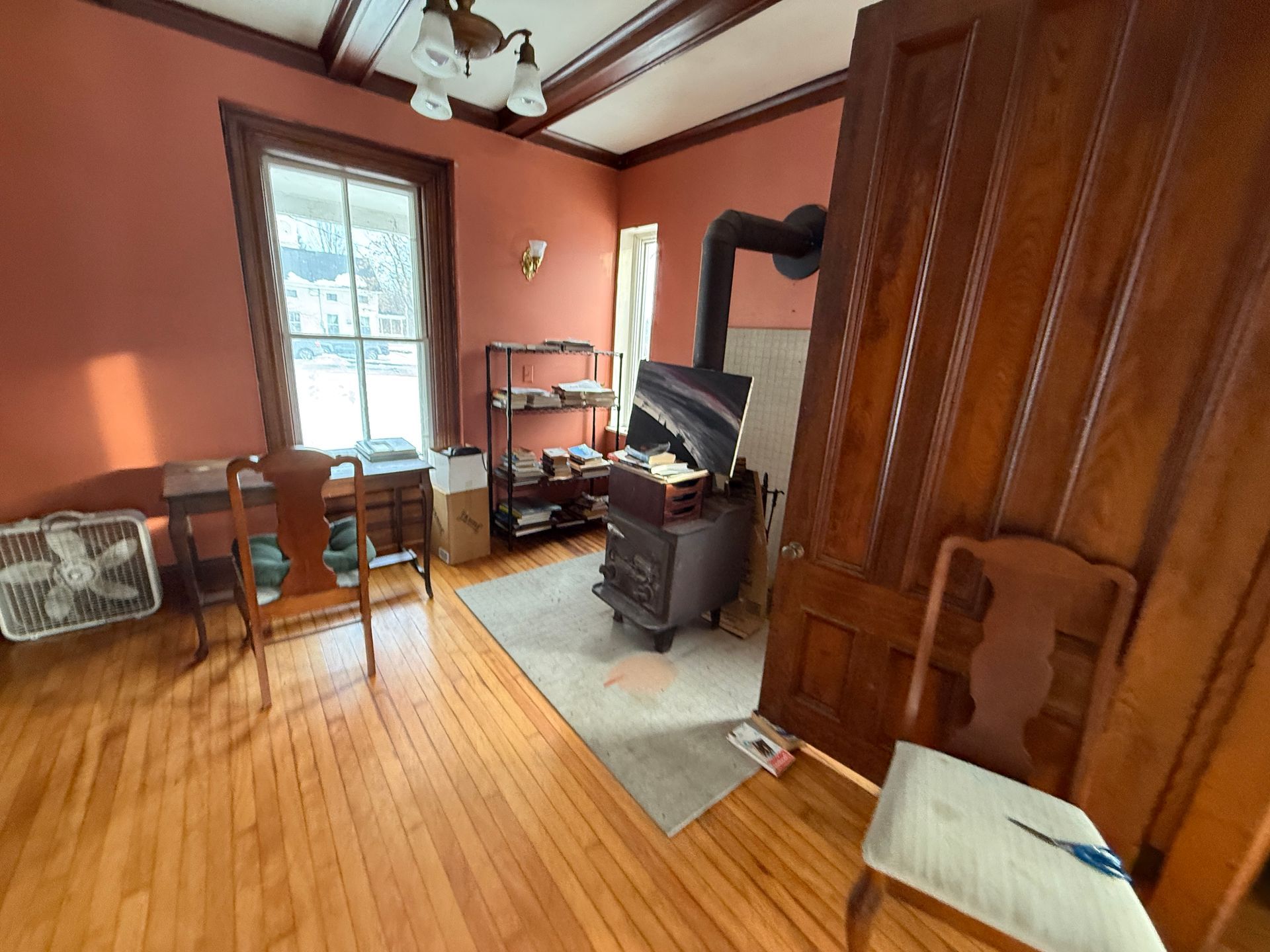 Room with wooden floor, wood-burning stove, desk, bookshelves, and window. Pink walls, brown door, and ceiling beams.