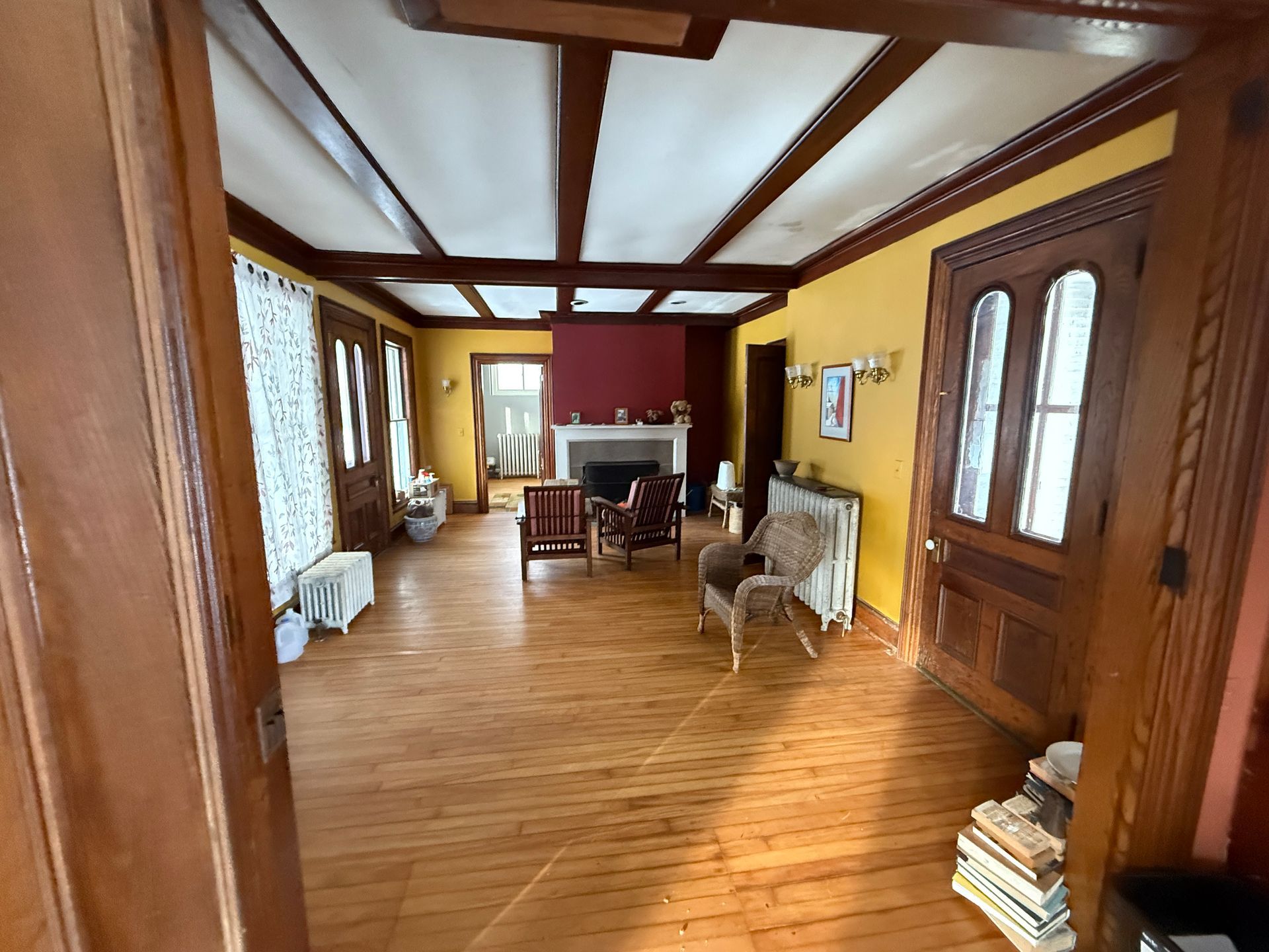 Spacious room with wood beams, yellow walls, and fireplace. Wooden door and floor with chairs, curtains, and books.
