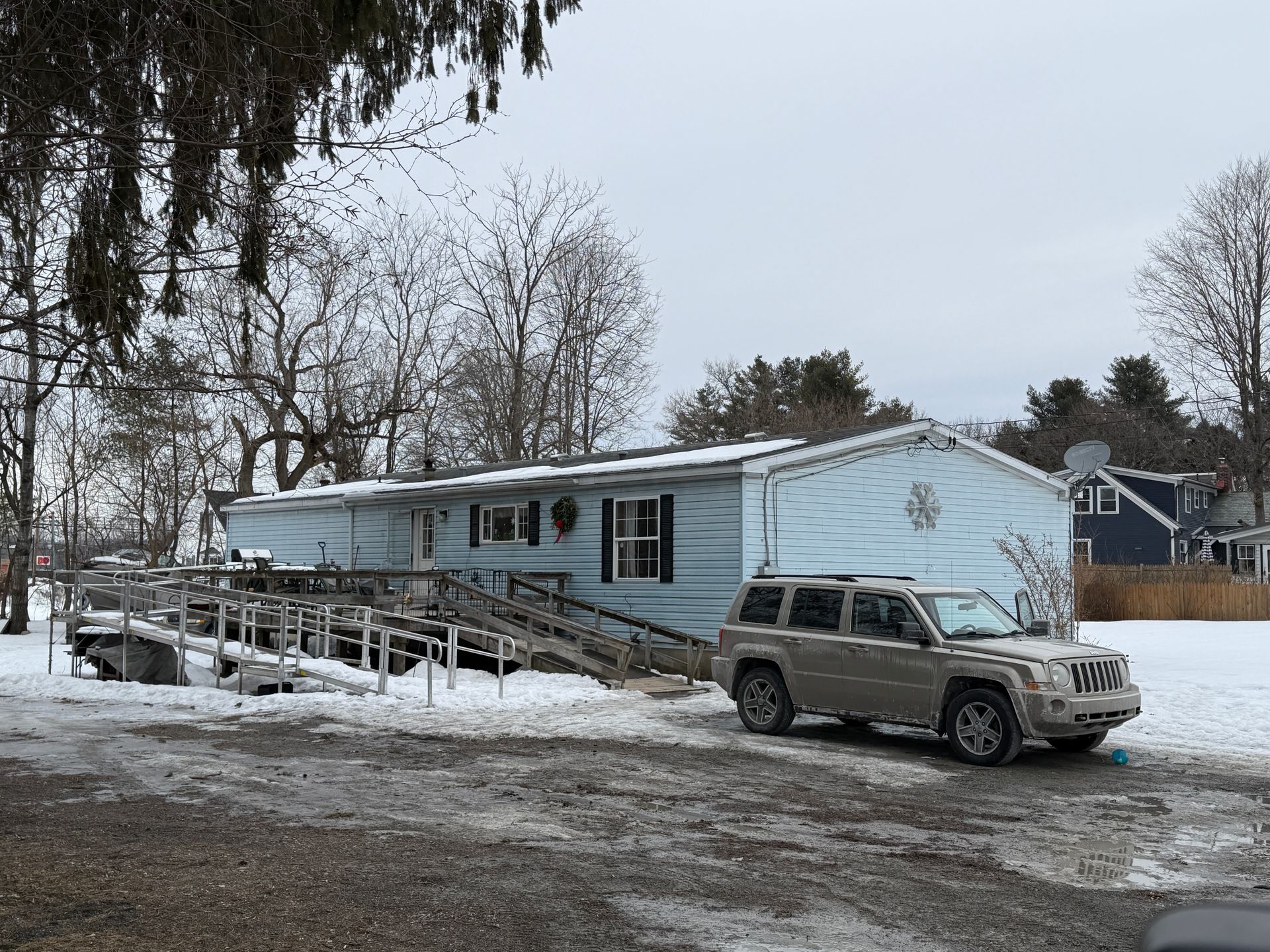 Light blue mobile home with a wooden ramp, a beige SUV parked in front, snow-covered ground.