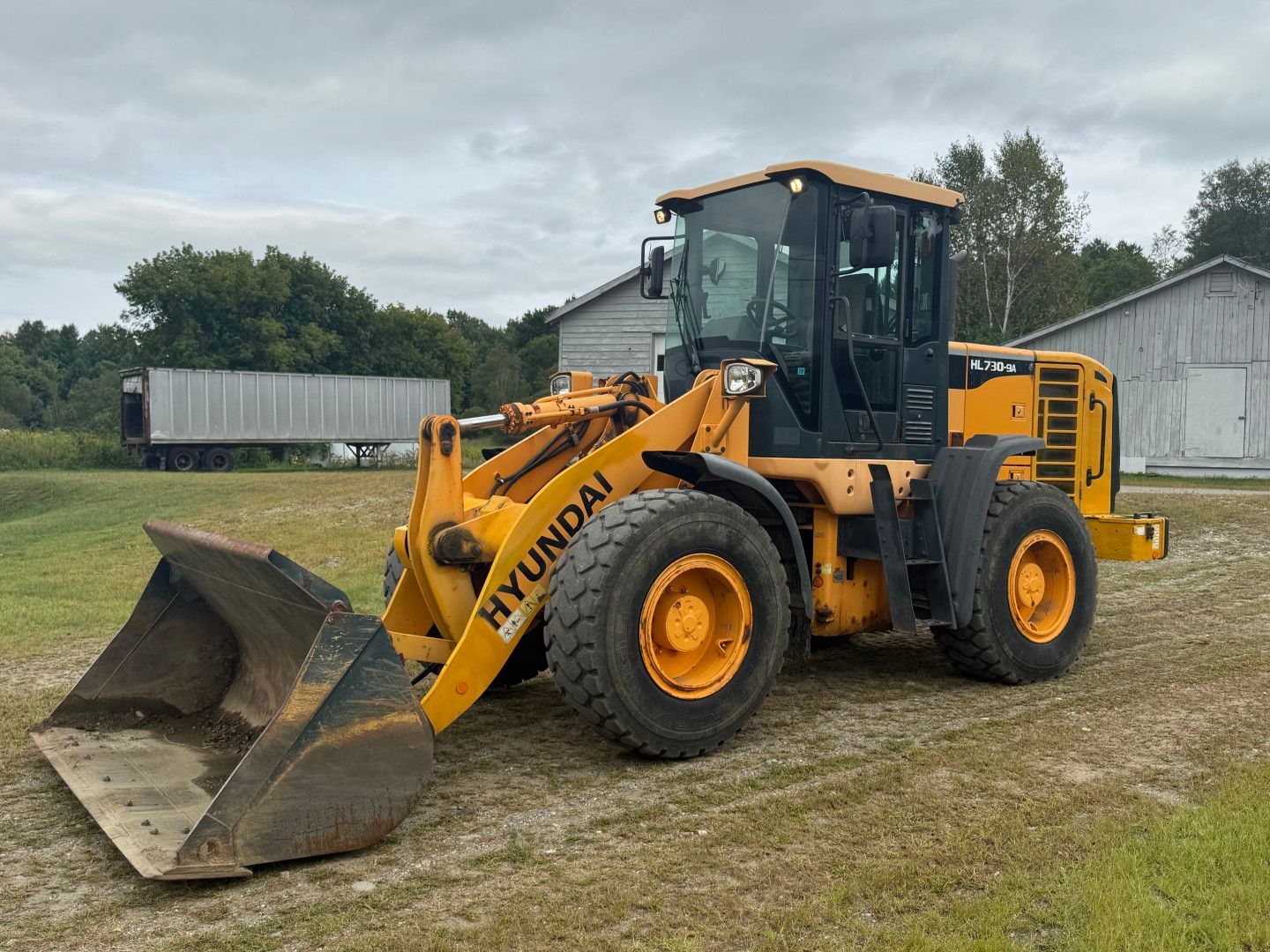 Yellow front loader on a grassy lot with a large bucket, buildings, and a cloudy sky in the background