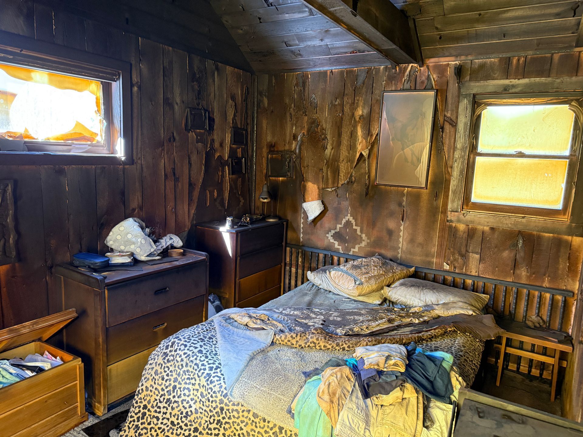 Interior of a room with fire damage, with two windows, a bed, dressers, and a small wooden chest.