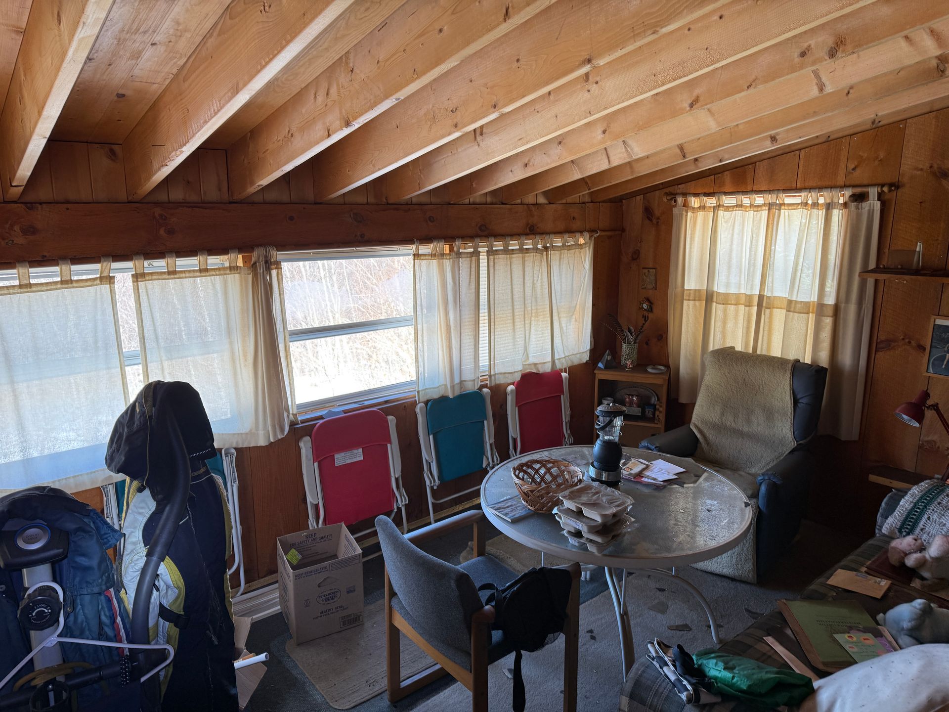 Interior of a small cabin. Windows with curtains, wood ceiling, round table with items, several chairs, and a box.