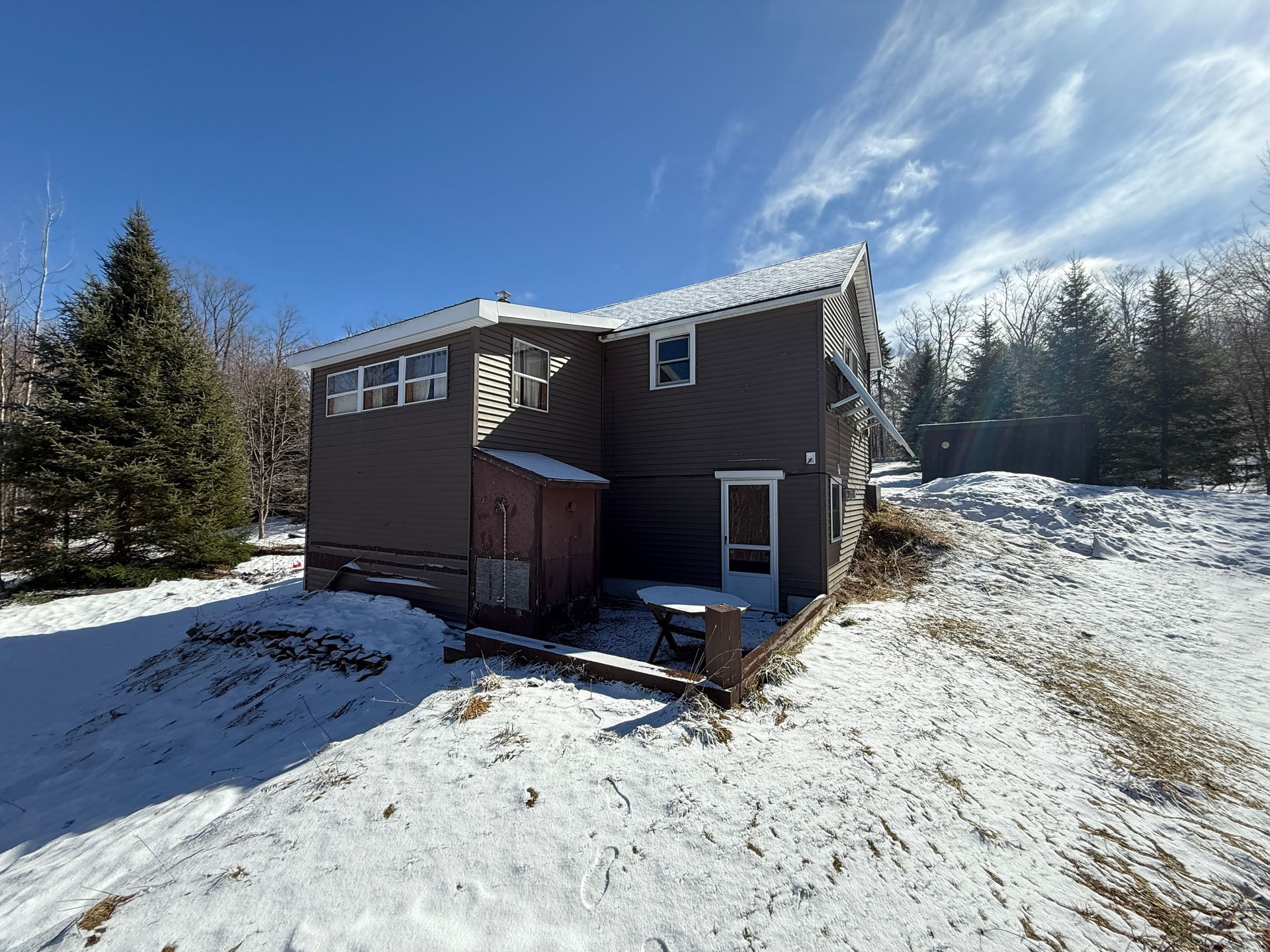 Snow-covered brown house with small shed, table, and tall trees under a partly cloudy blue sky.