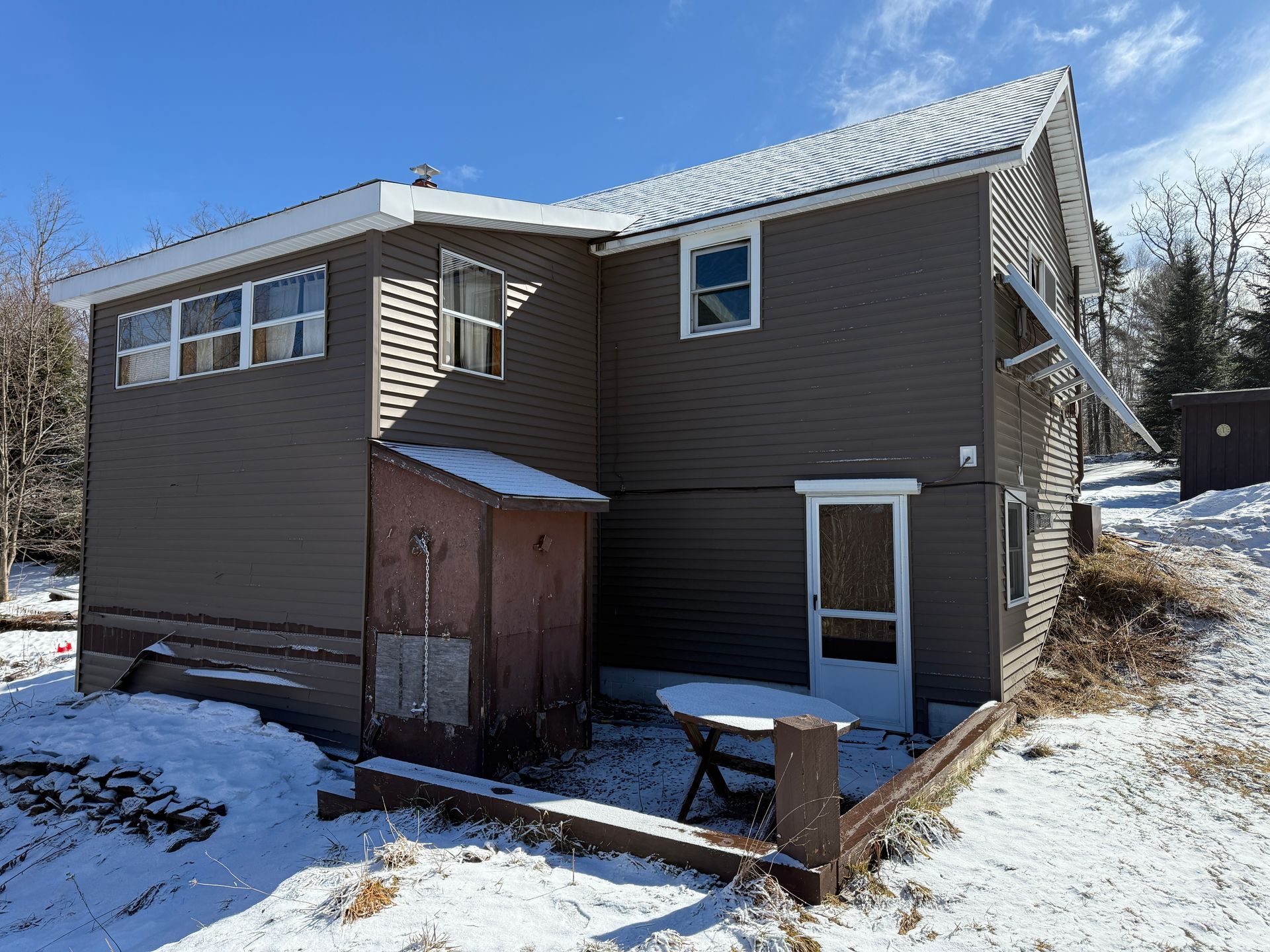 Brown two-story house with snowy roof and ground. Small shed and white door visible. Clear blue sky.