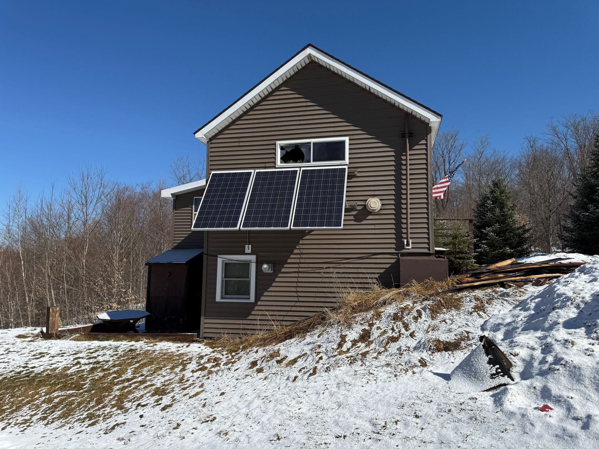 Brown cabin with solar panels, snow-covered ground, clear blue sky, and American flag.