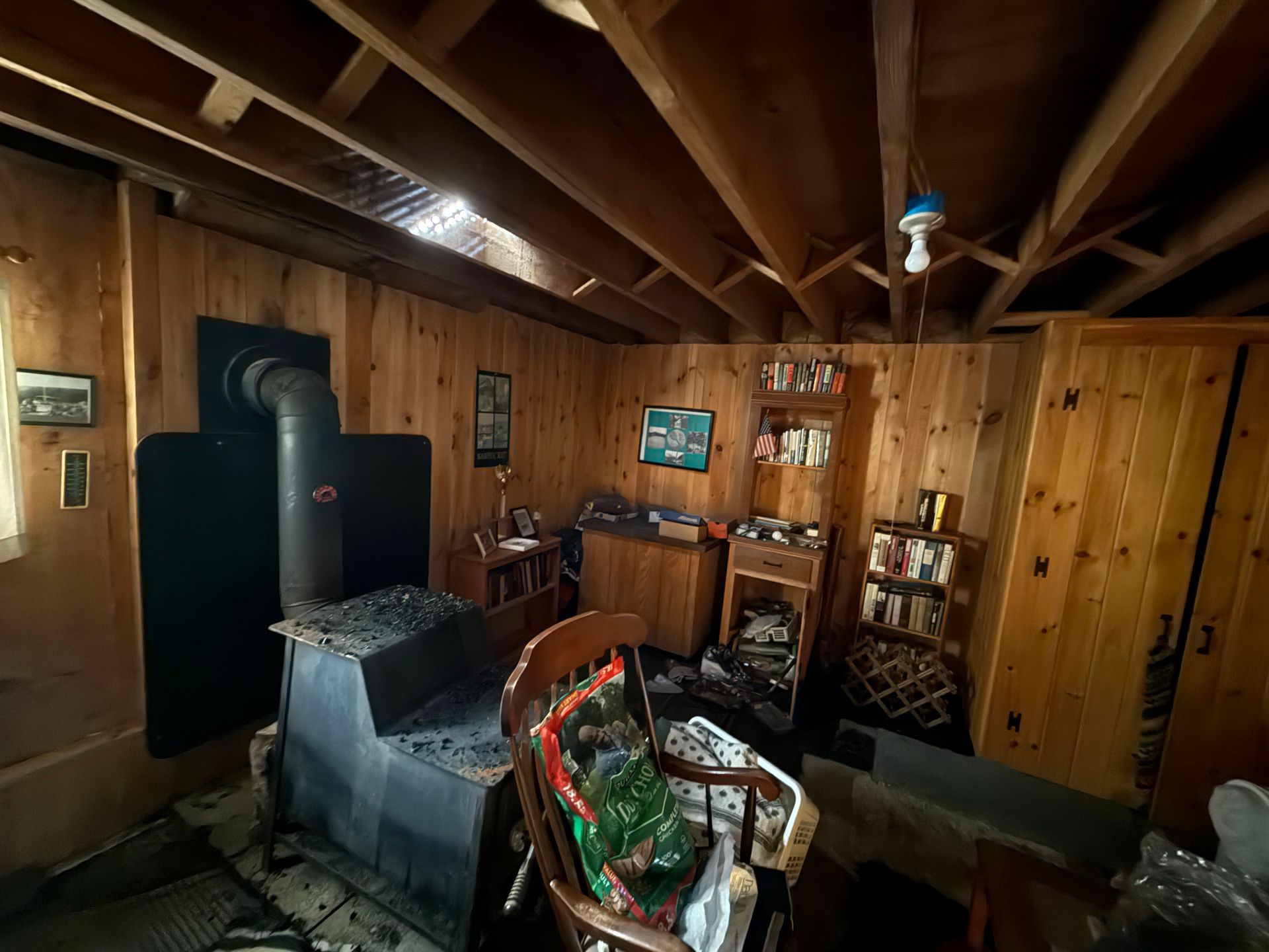 Interior of a cabin with a wood-burning stove, wooden walls, a small bookshelf, and a chair.