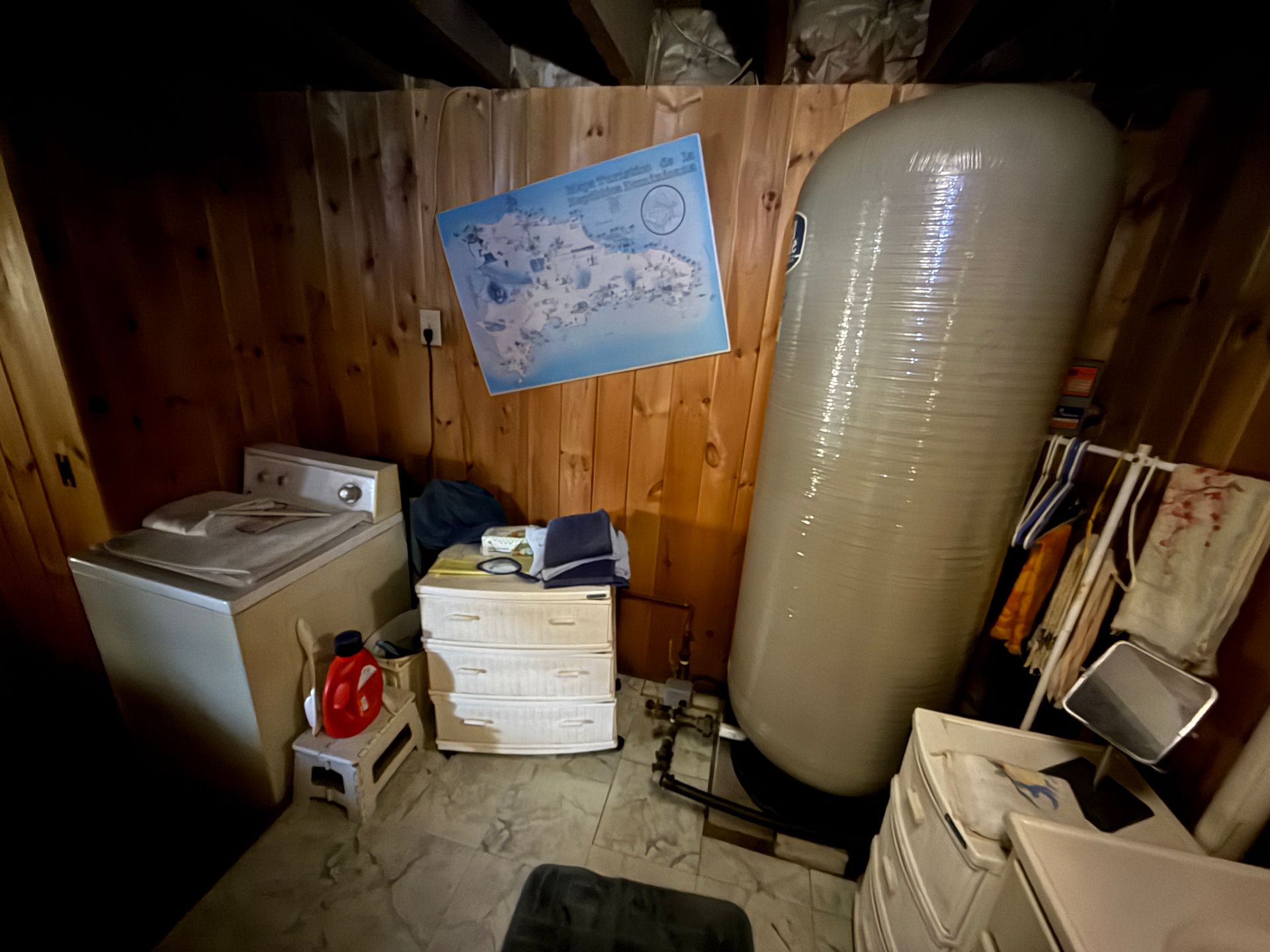 Laundry room with a washing machine, water tank, and a dresser against wood-paneled walls.