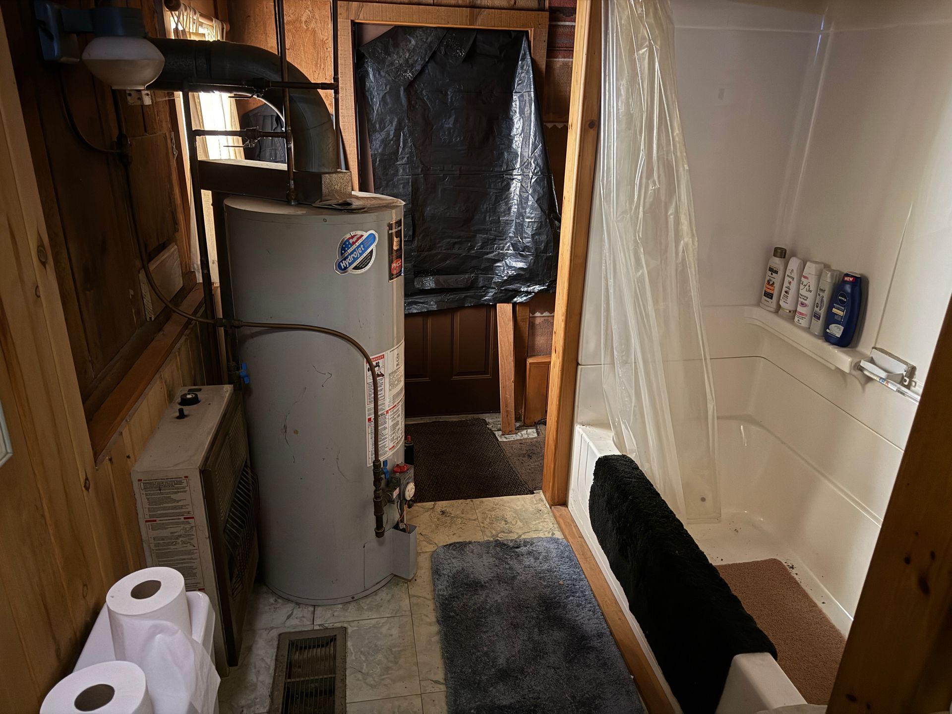 Bathroom with water heater and shower. Wooden walls, gray rug, and toilet paper.