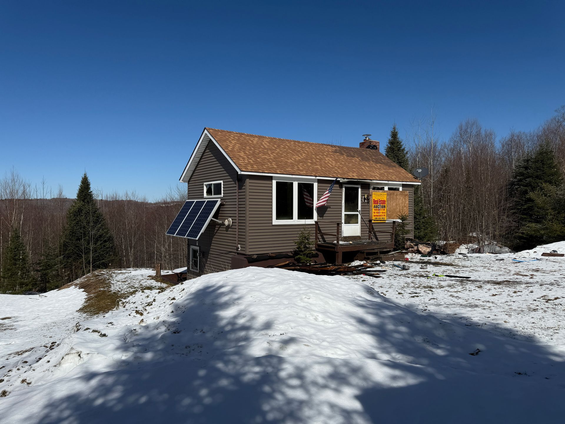 Small cabin with solar panel, snowy ground, and trees under a clear blue sky.