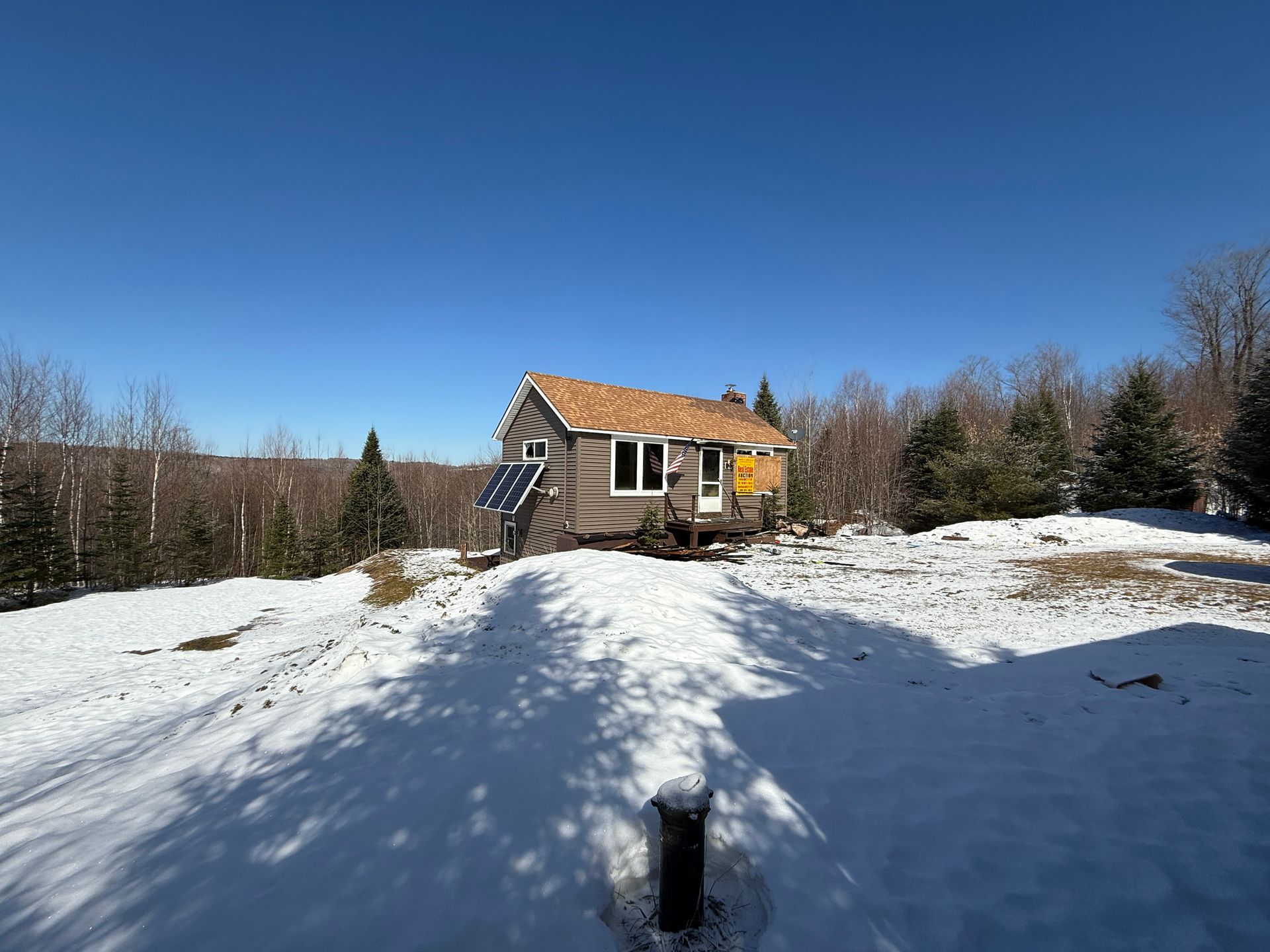 Snow-covered landscape with small cabin under a bright blue sky. Solar panel and bare trees.