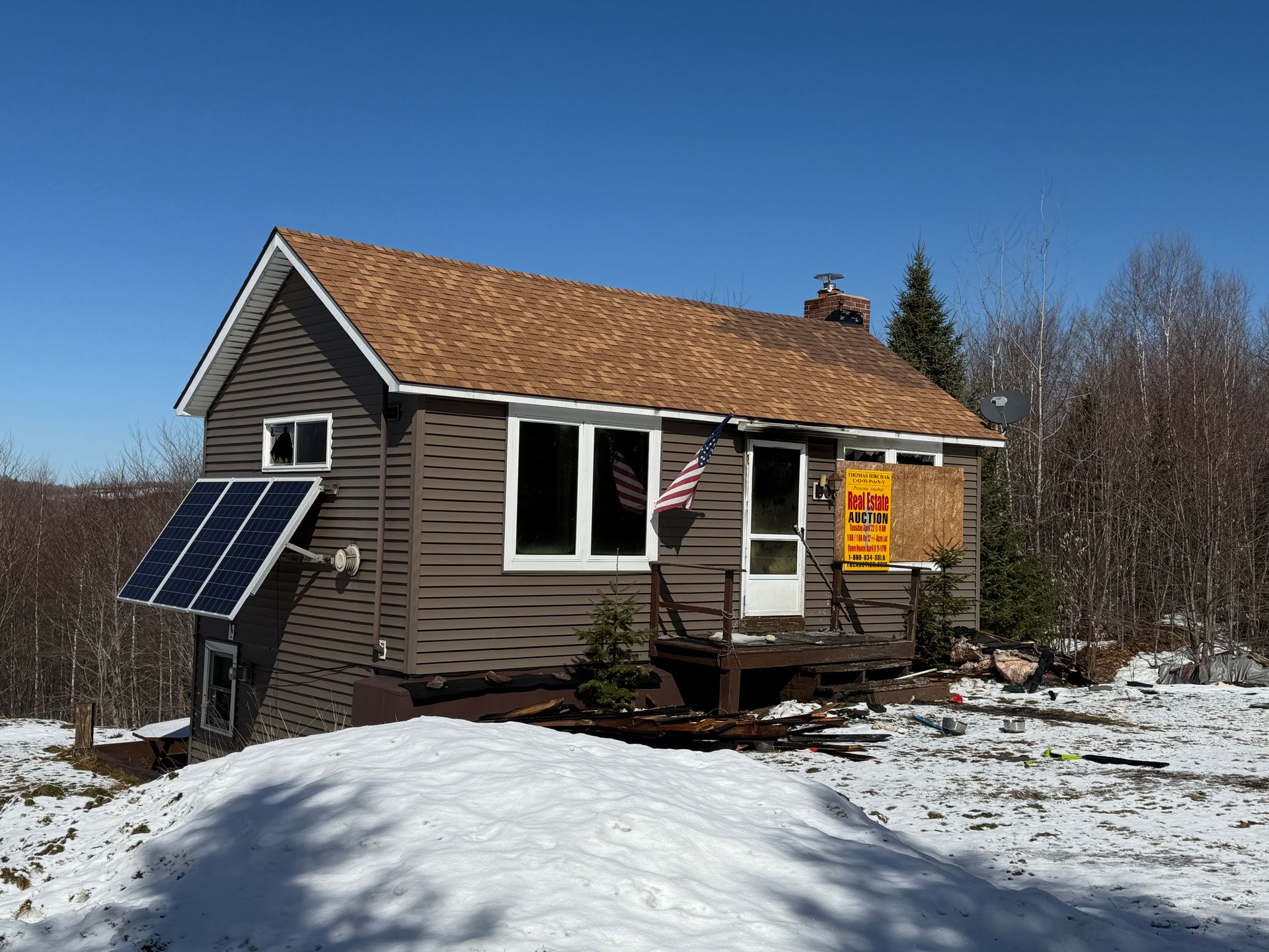Small, brown cabin with solar panel in snowy setting, American flag, clear blue sky.
