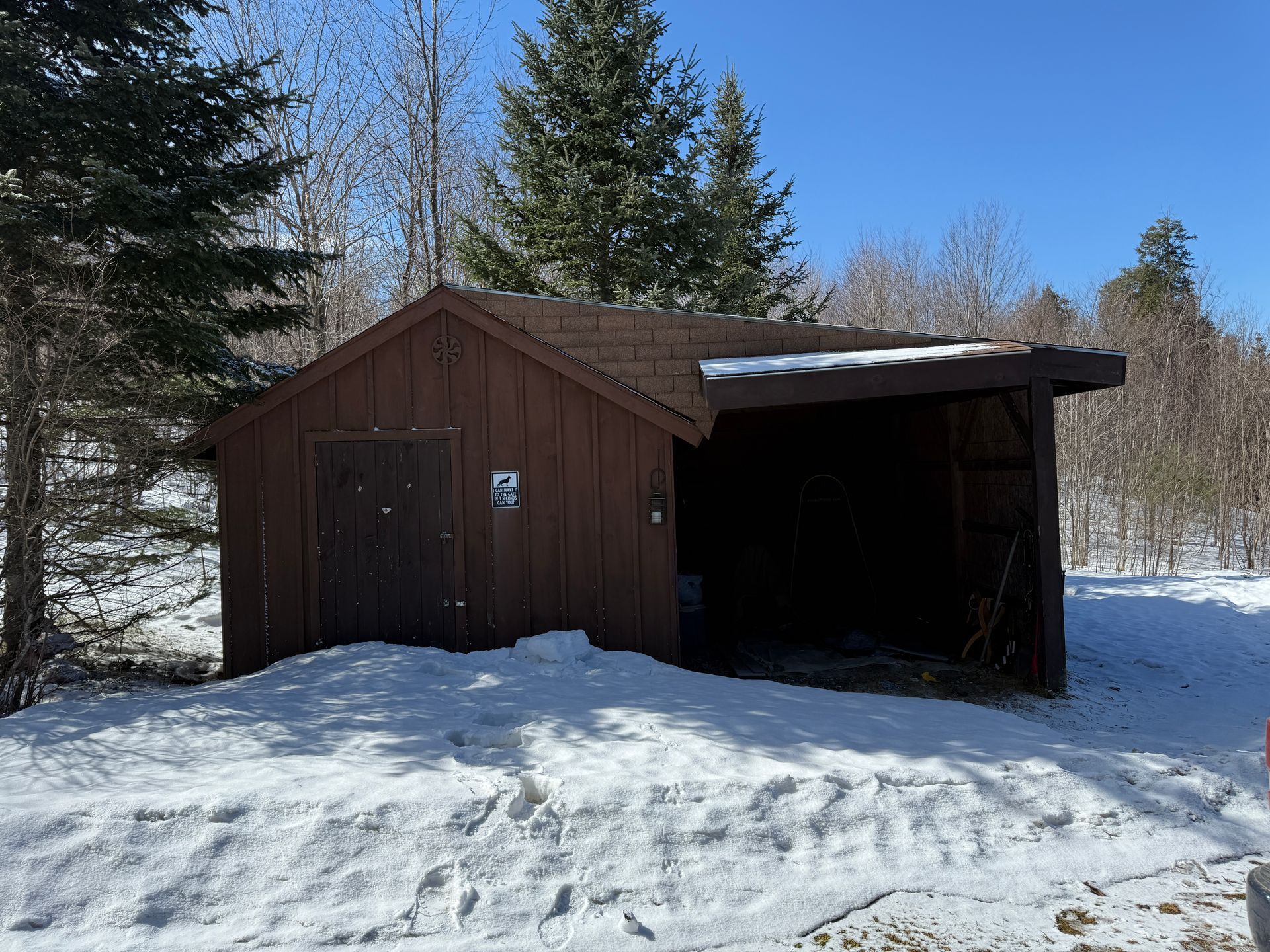 Brown shed with closed door and open side, surrounded by snow, trees, and blue sky.
