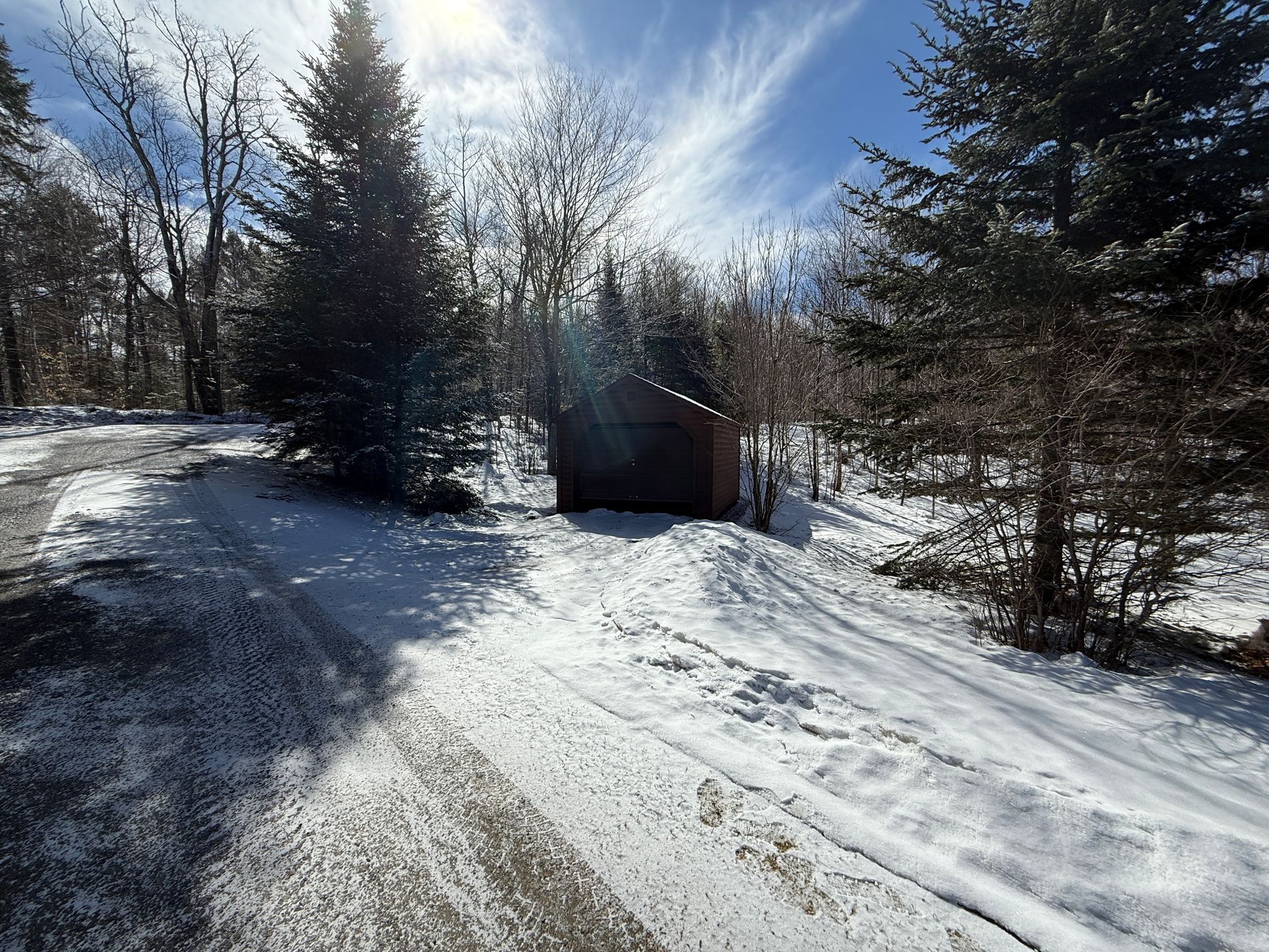 Snowy road leading to a small, dark wooden structure among evergreen trees on a sunny day.