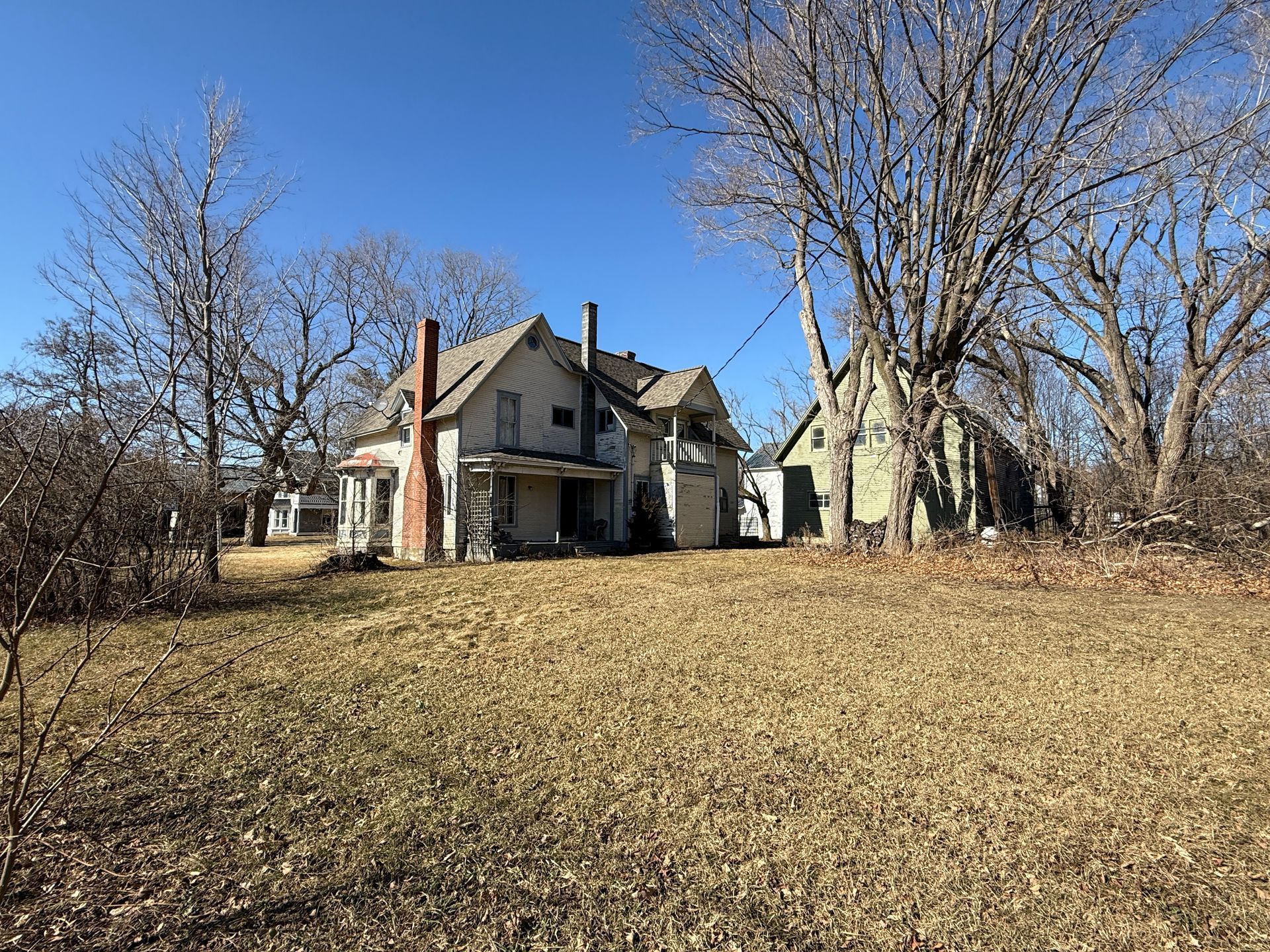 House and barn on a grassy lot under a clear, blue sky. Bare trees surround the buildings.