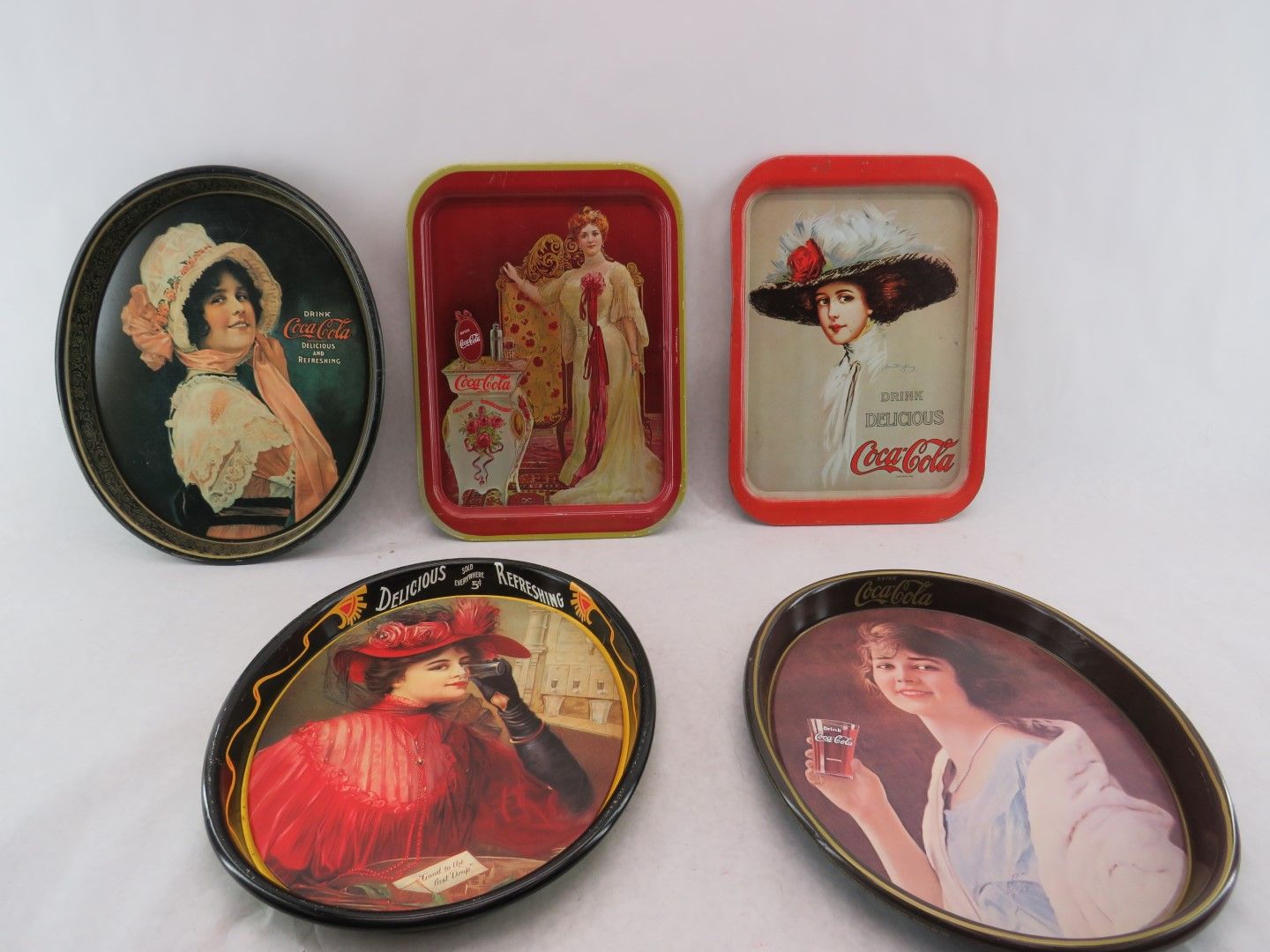 Five vintage Coca-Cola serving trays, featuring women in antique clothing; various shapes and colors.