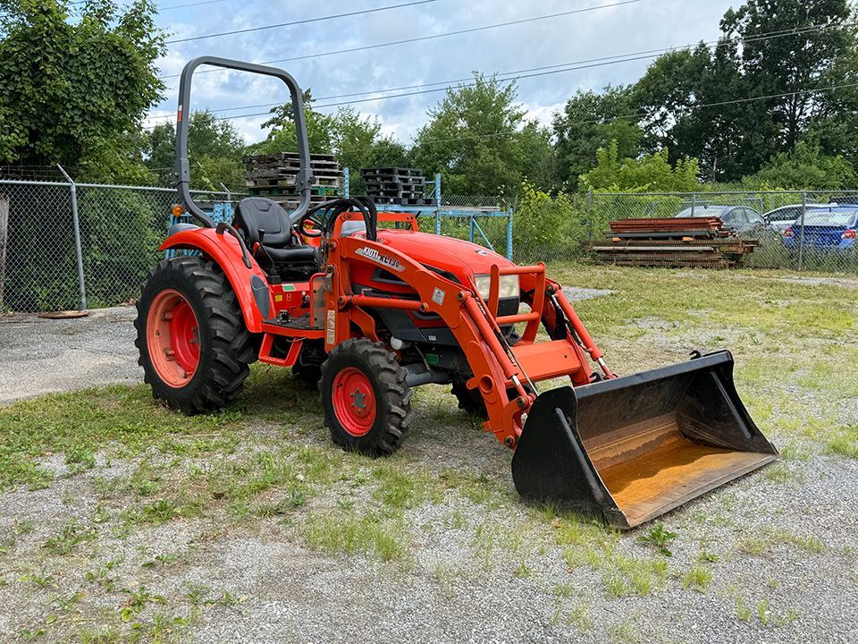 Orange tractor with front loader parked on gravel near a chain-link fence and trees