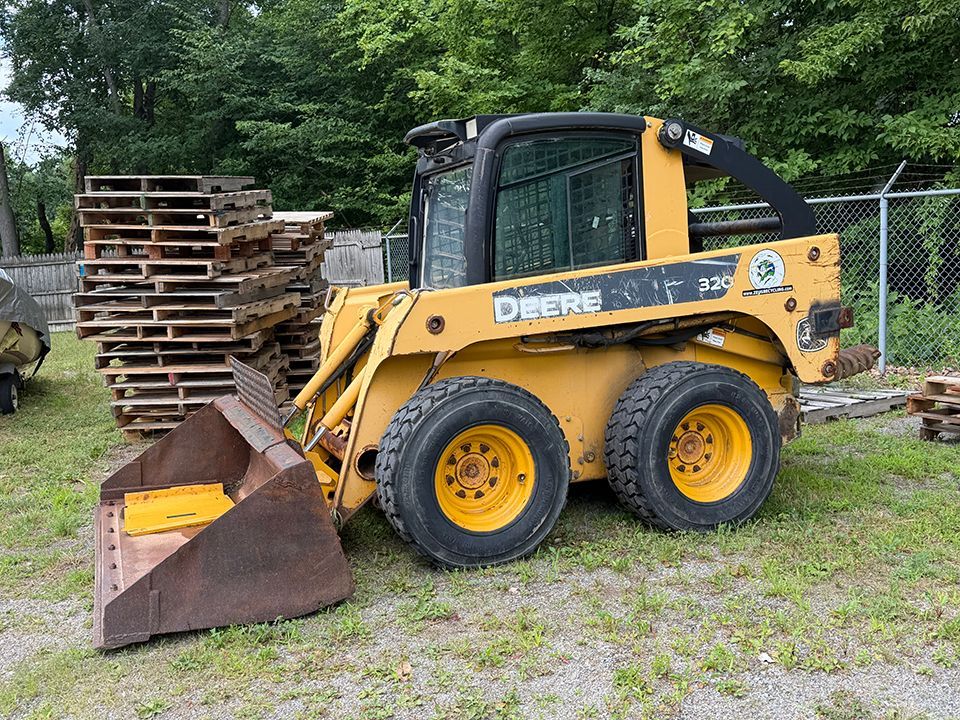 Yellow John Deere skid steer with front bucket parked on gravel beside stacked wooden pallets.