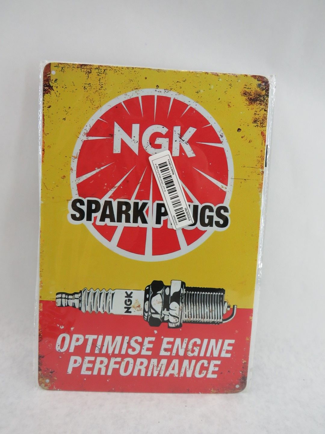 Metal sign for NGK spark plugs; yellow, red, white colors, distressed look, text and image of a spark plug.