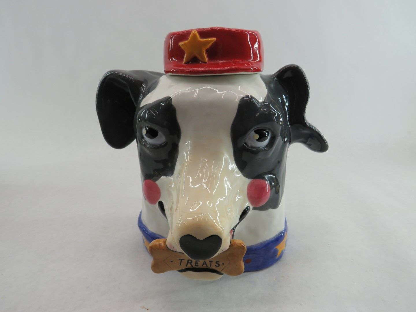 Dog-shaped ceramic treat jar. Black and white face, red hat with gold star, holding bone-shaped treat that reads “Treats.”