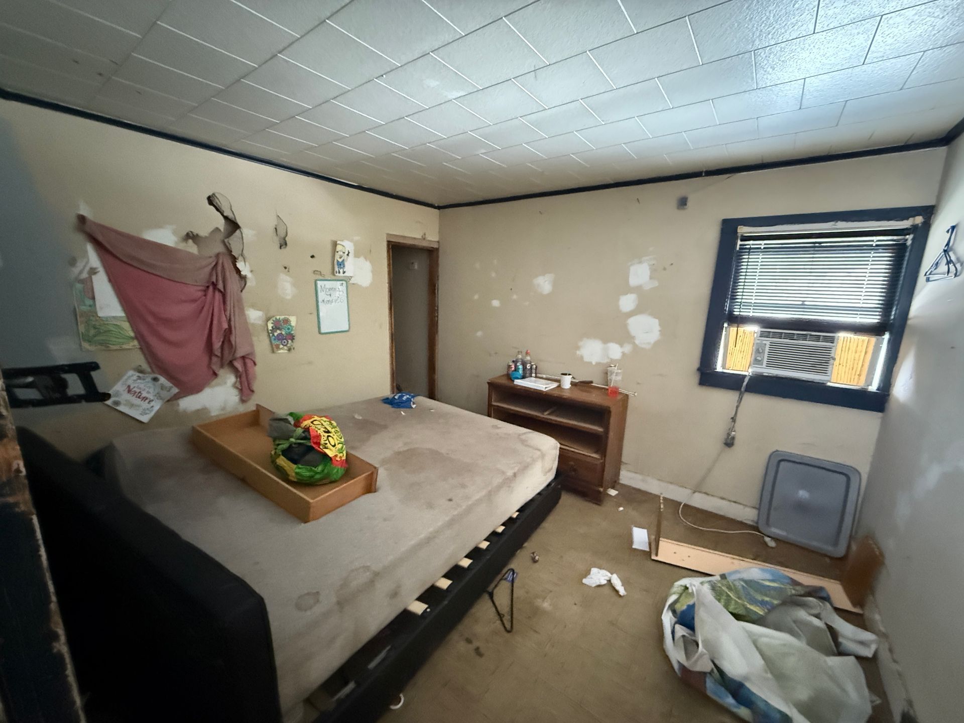 A bedroom with a mattress on a black bed frame, a small wooden dresser, and a window with an AC unit.