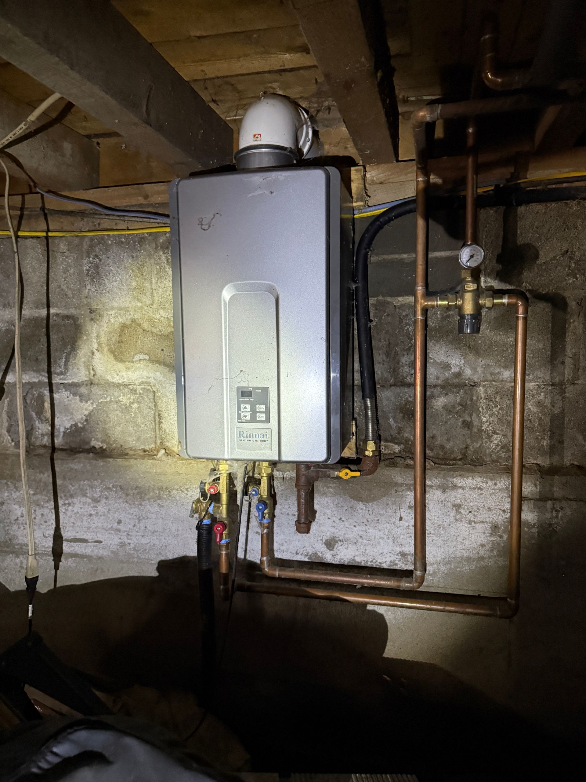 A silver tankless water heater mounted on a concrete wall in a basement, connected to copper pipes and a white vent pipe.