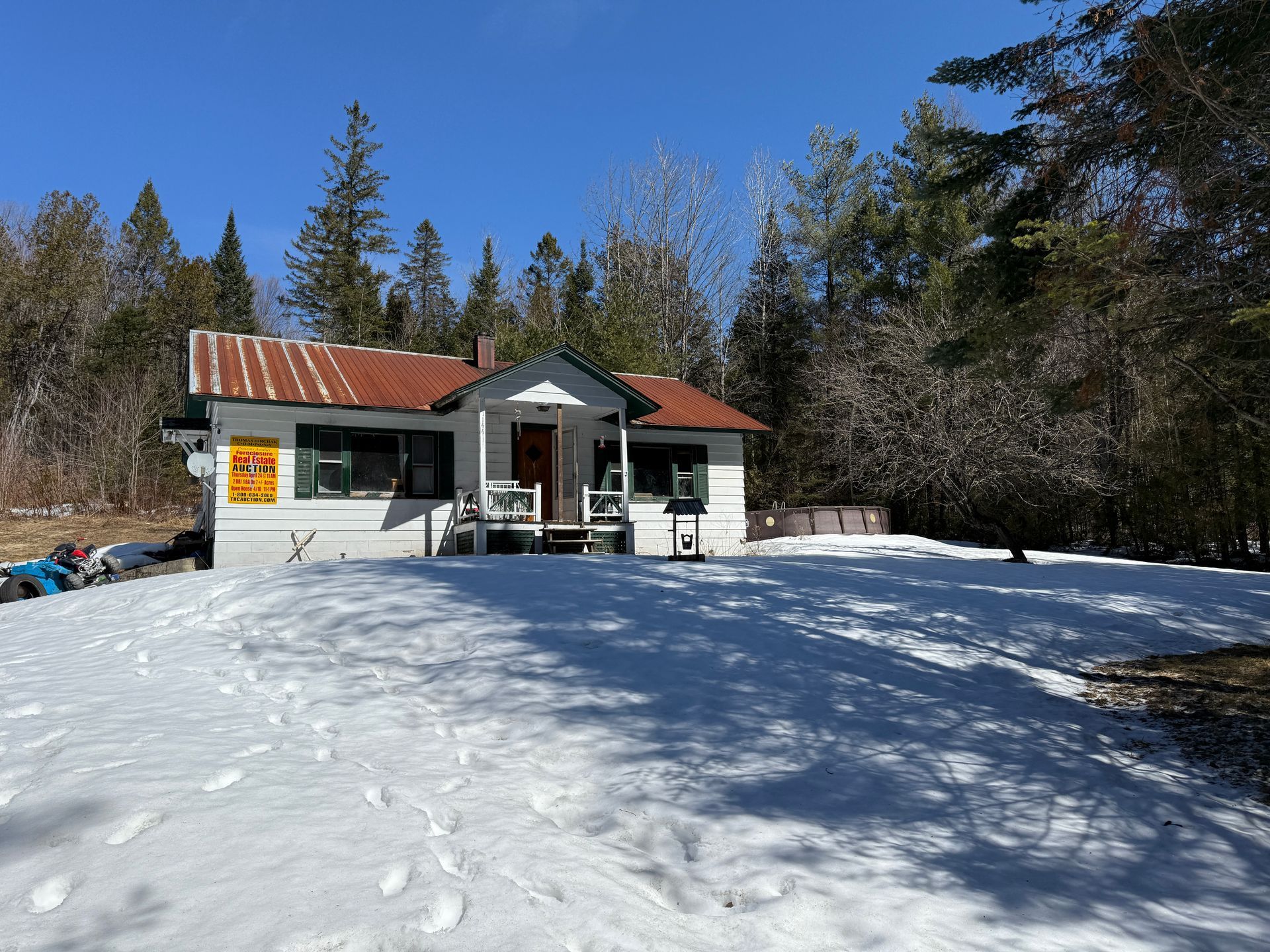 A white cabin with a metal roof stands in a snow-covered clearing, surrounded by dense evergreen trees.