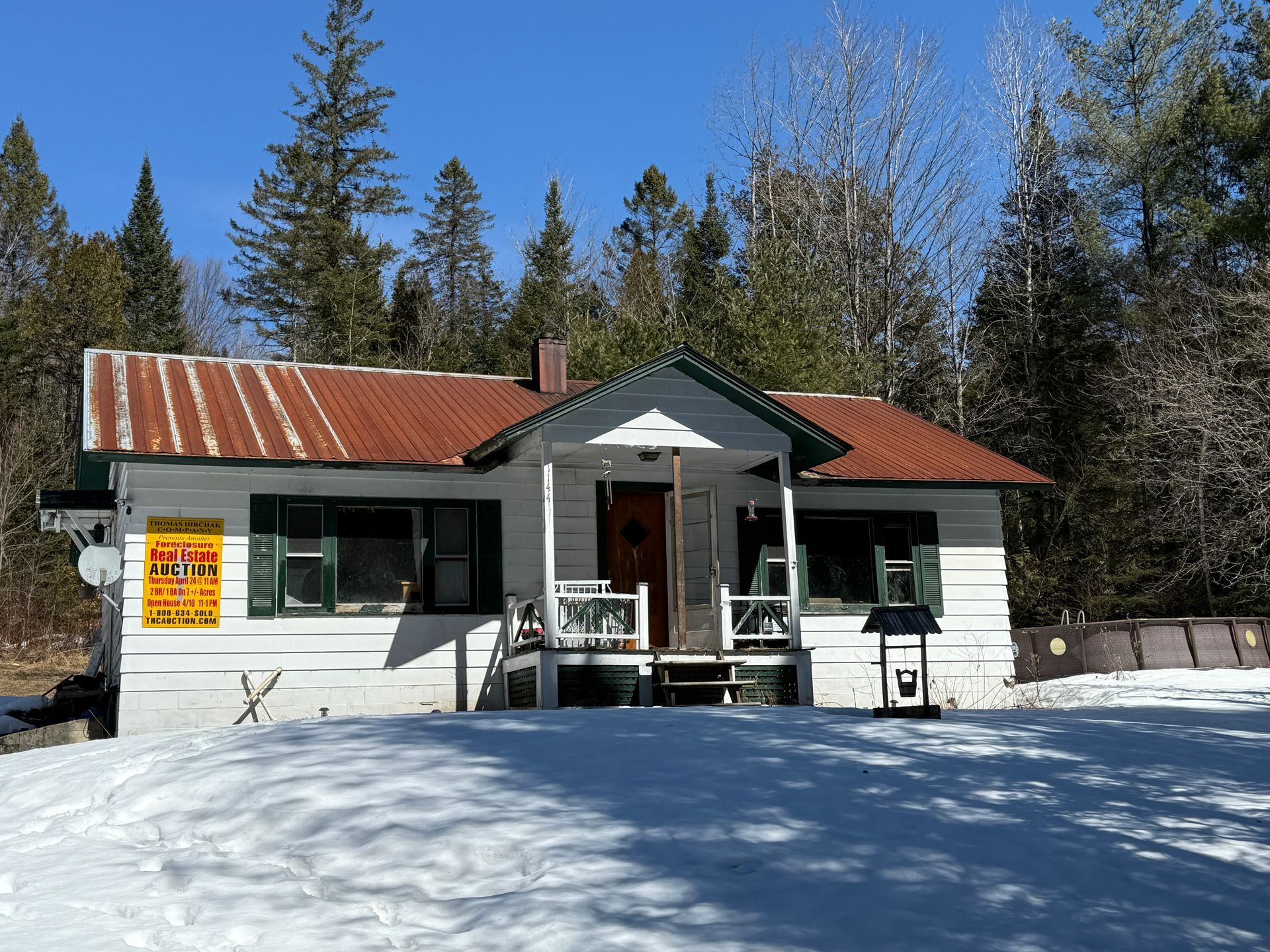 A white cabin with a red tin roof sits in a snowy clearing surrounded by a dense forest under a clear blue sky.