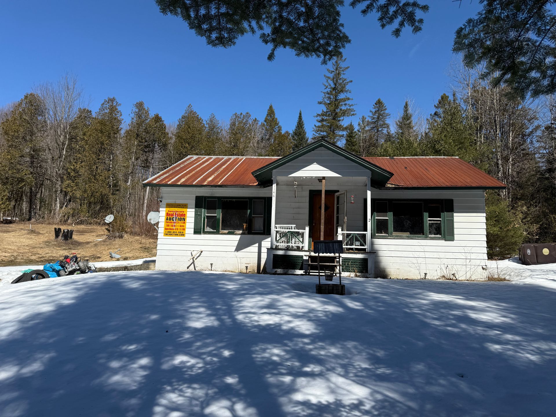 A white, one-story cottage with a metal roof, standing in a snowy clearing in front of a forest of pine trees.