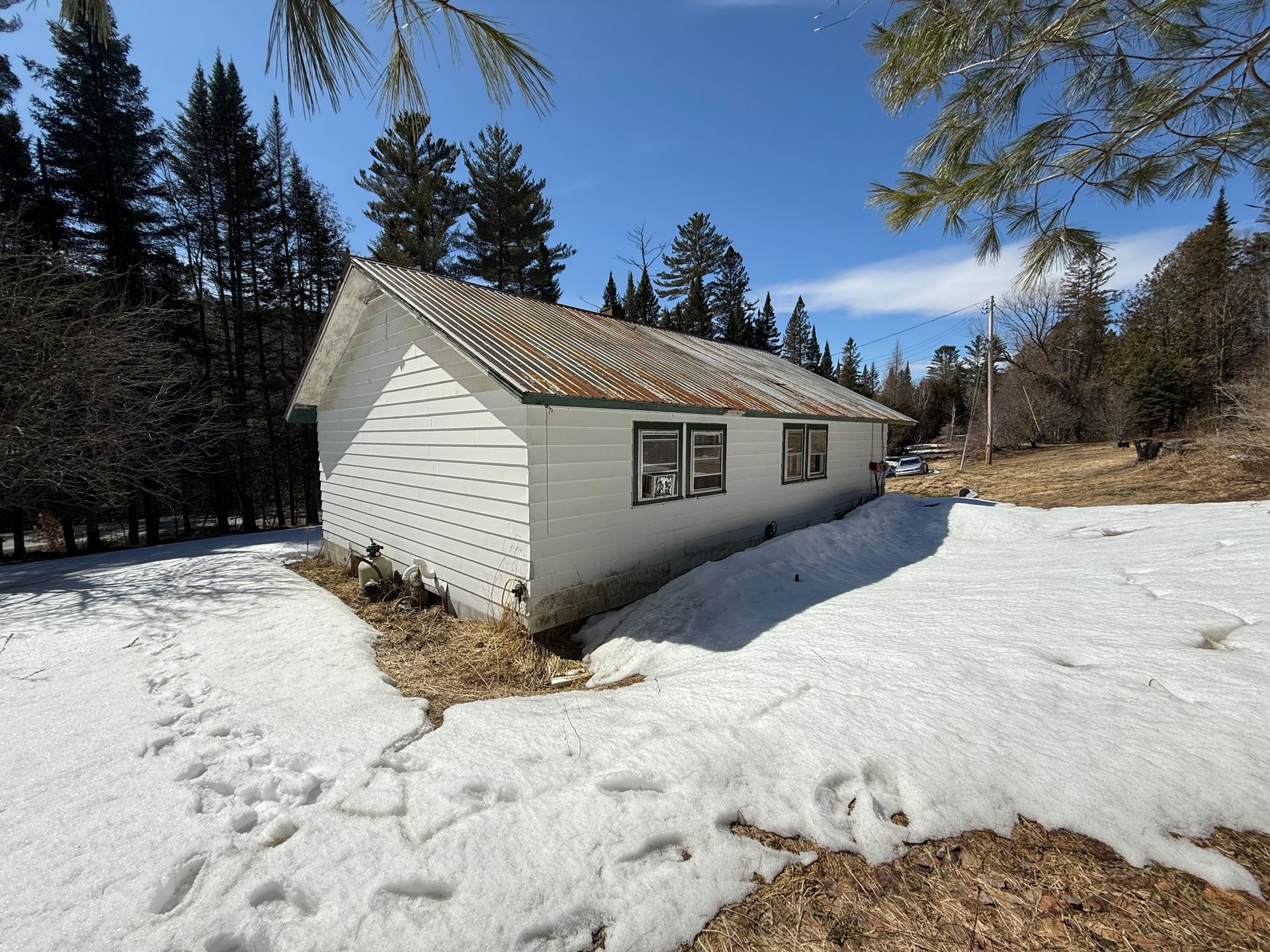 A white, single-story cabin with a brown shingled roof sits in a snowy clearing surrounded by pine trees under a blue sky.