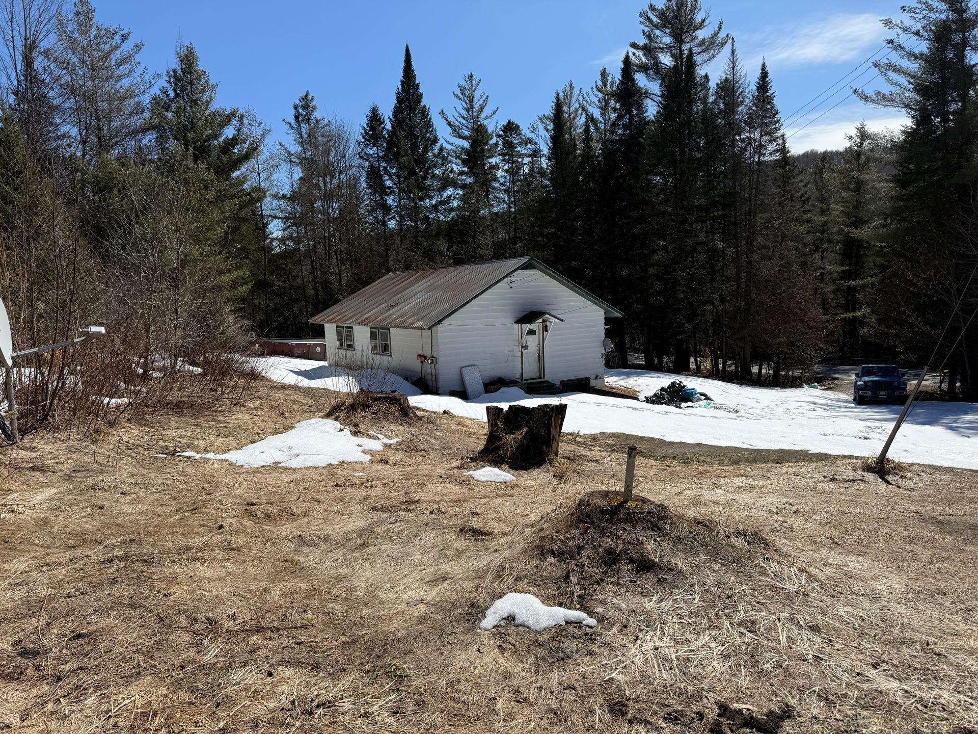 A small, white house with a dark roof sits in a wooded area with patches of snow on the ground under a clear blue sky.
