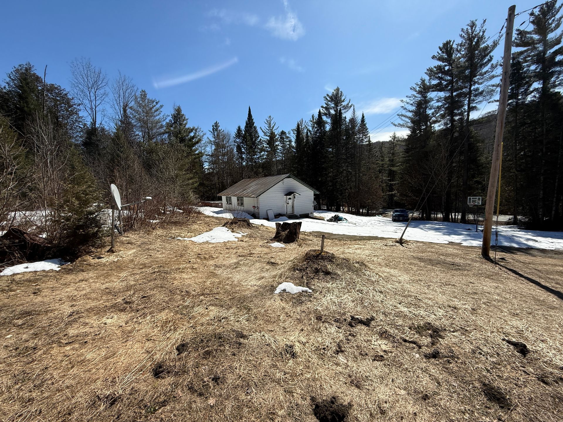 A white house sits in a clearing surrounded by evergreen trees, with patches of melting snow on the ground under a blue sky.