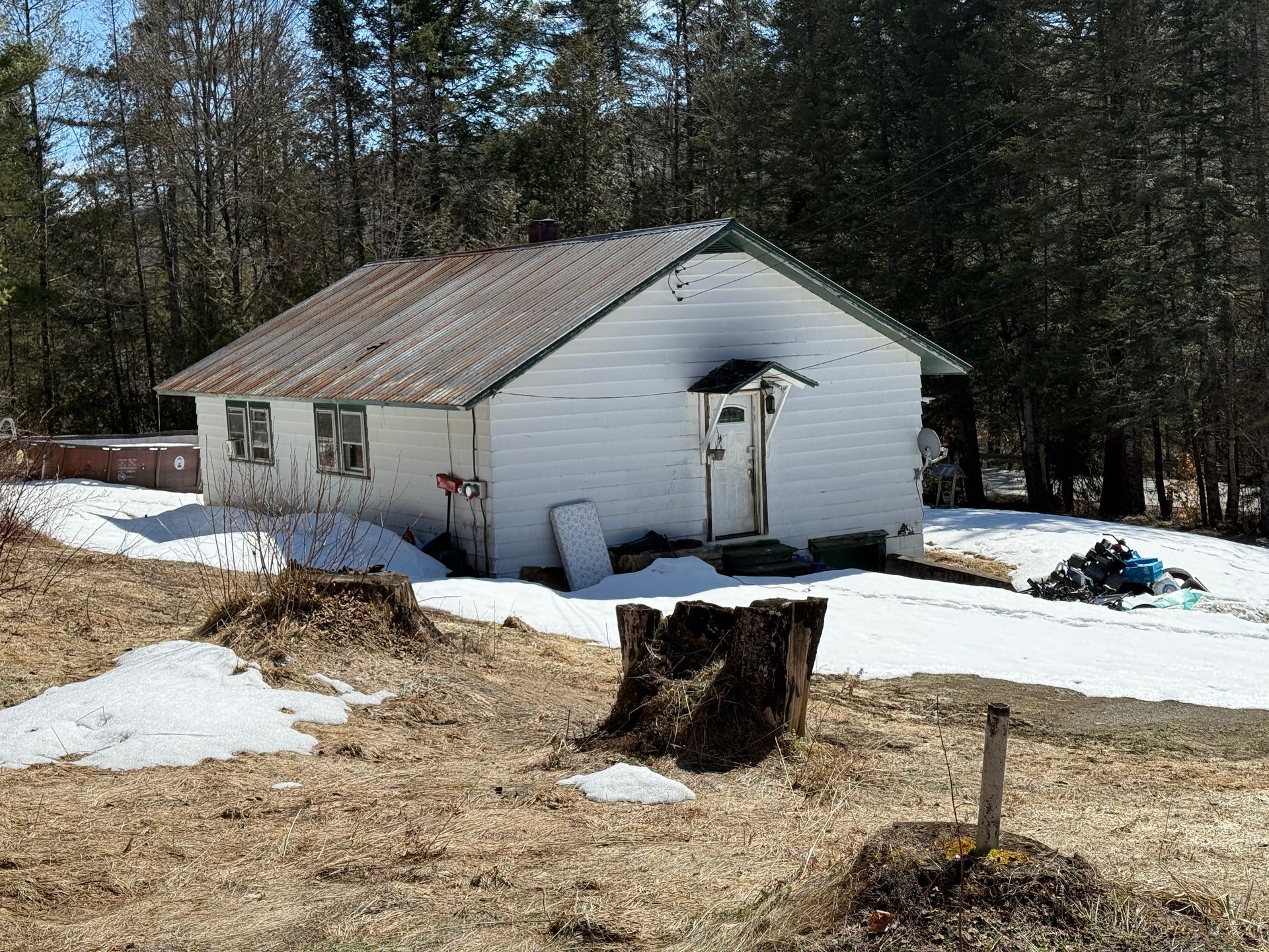 A white, single-story house with a gabled metal roof sits in a landscape with patchy snow and a tree stump in the foreground.